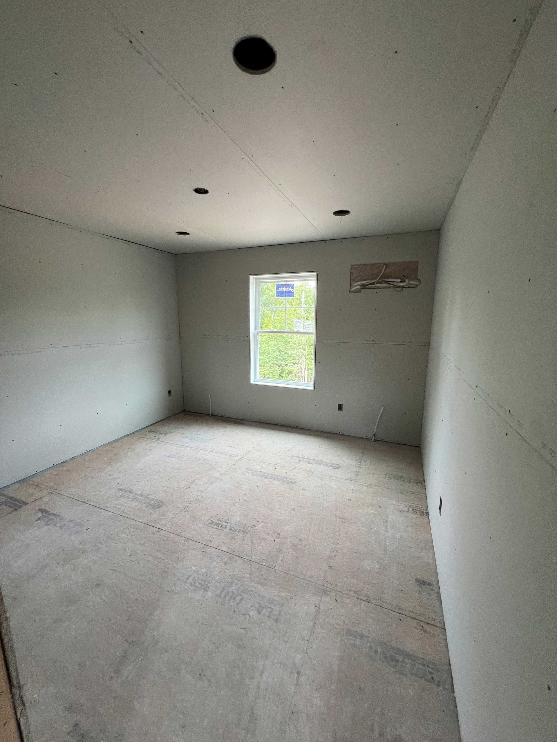 Empty room with unfinished drywall and concrete floor, single window, recessed lighting.