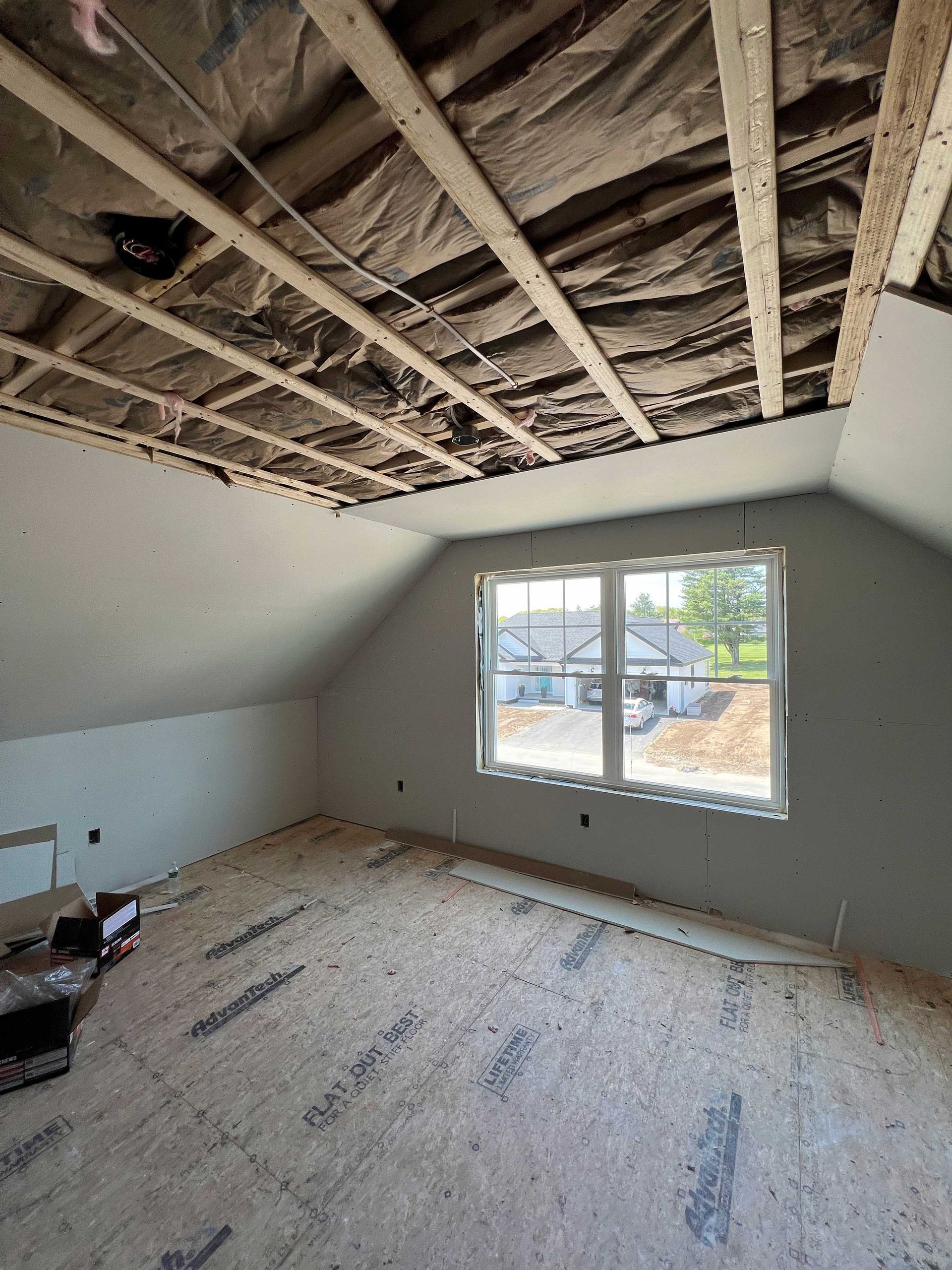 Interior of a room under construction, with a window, exposed ceiling beams, and unfinished flooring.