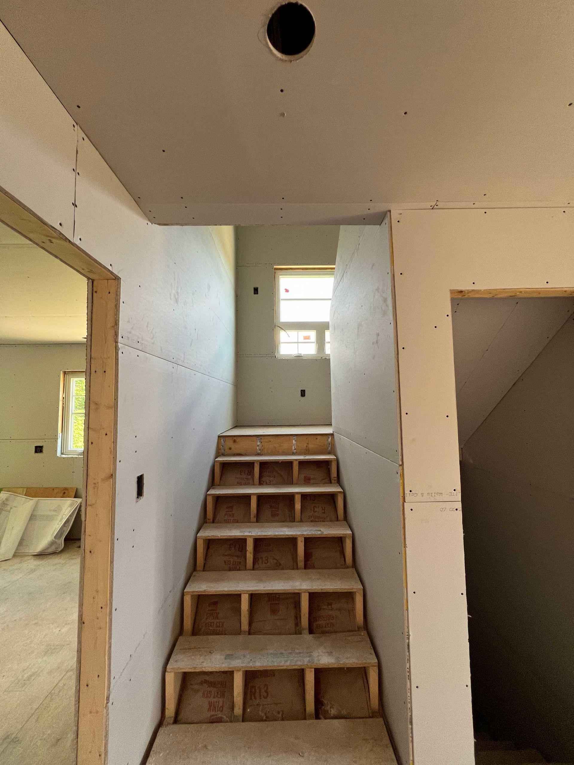 Staircase under construction with drywall and unfinished steps, leading to a small window.