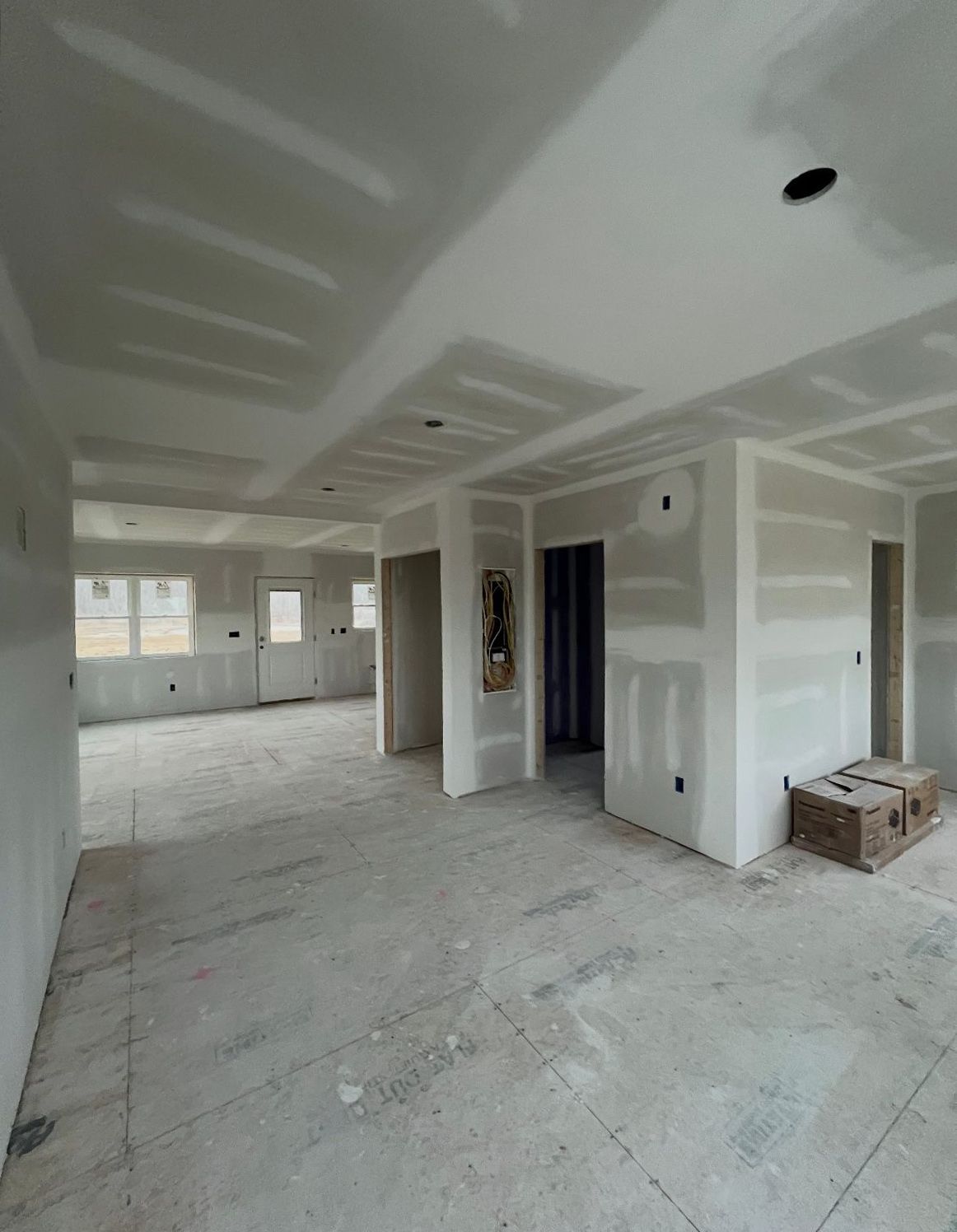 Interior of a house under construction; drywall and concrete floors are visible, with doorways leading to other rooms.
