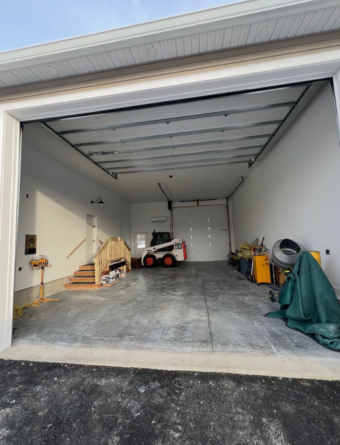 Open garage interior, concrete floor, white walls, small orange construction vehicle, tools.