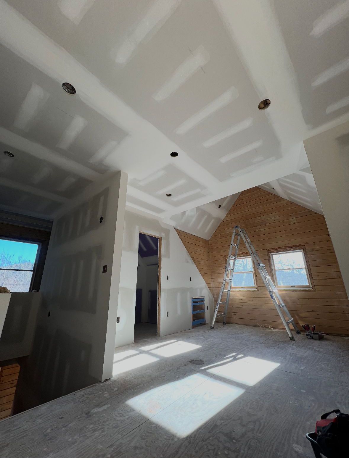 Interior under construction. Drywall, exposed brick, windows, and ladder visible in a room with angled ceilings.