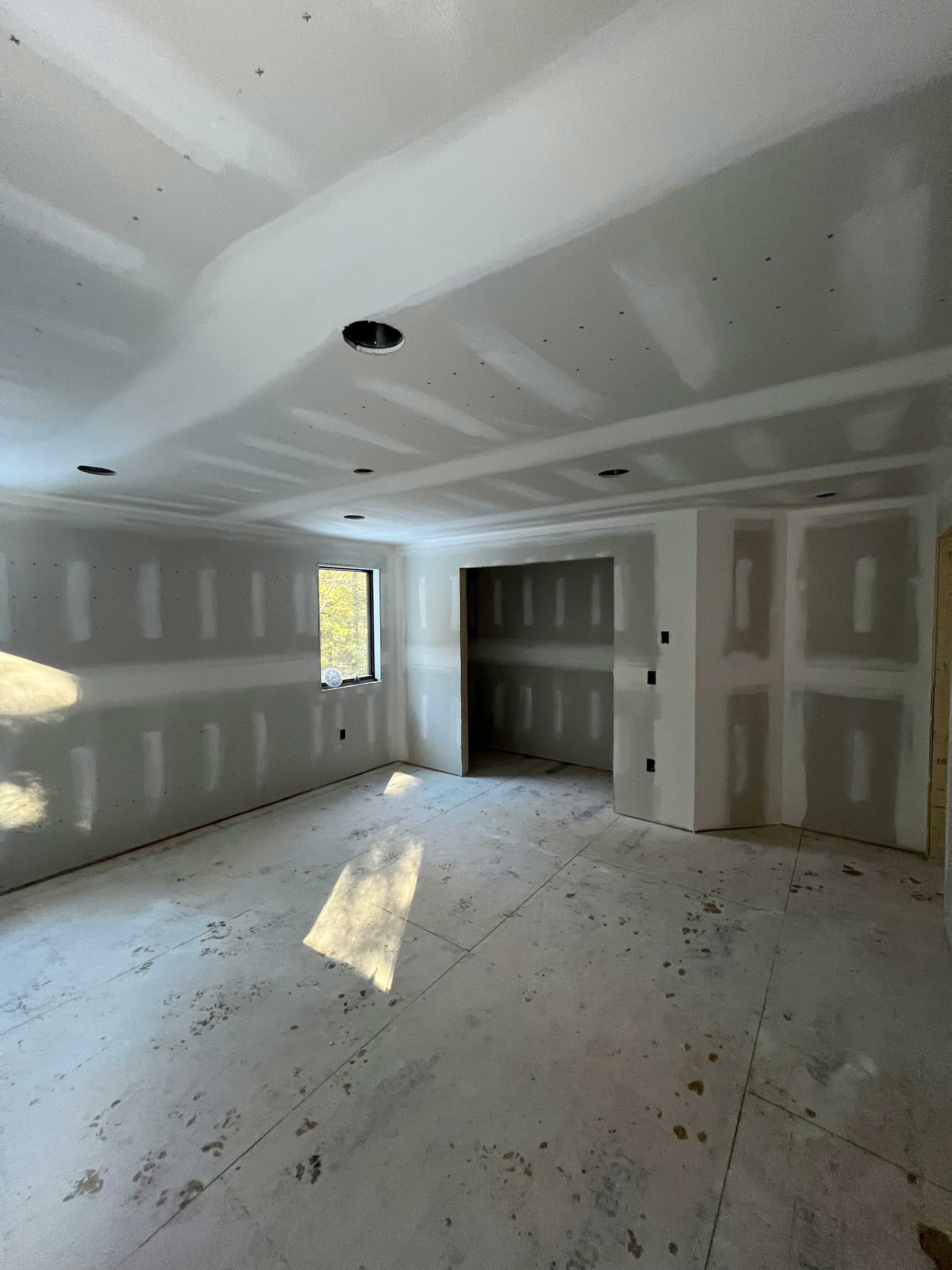 Interior view of unfinished room with drywall on walls and ceiling, light streaming in from a window.