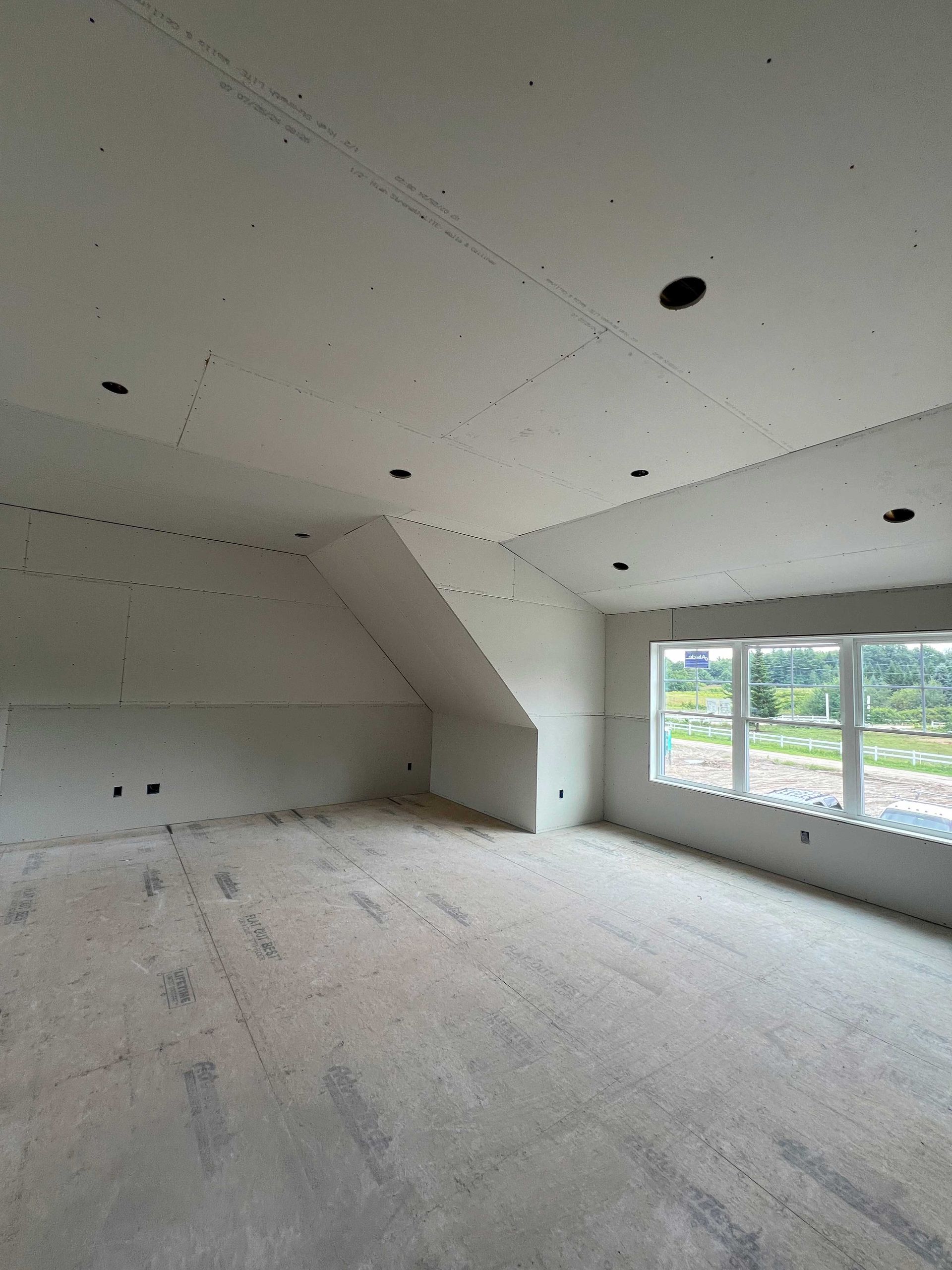 Interior view of a room under construction with drywall installed.  Unfinished walls, ceiling with recessed light openings, and a window.