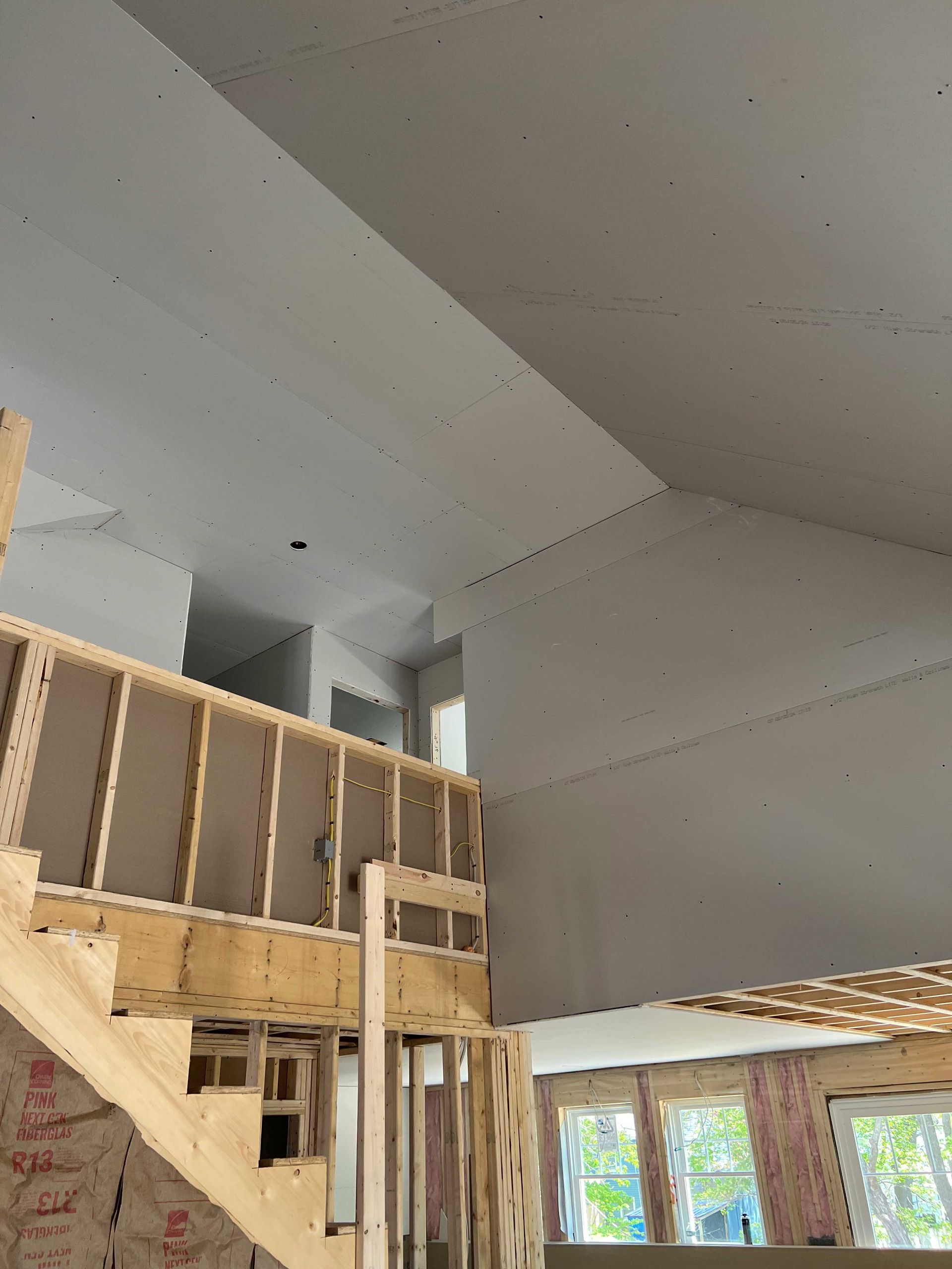 Interior of a house under construction. Drywall installed on the ceiling and walls, wooden framing visible on stairs and half-walls.