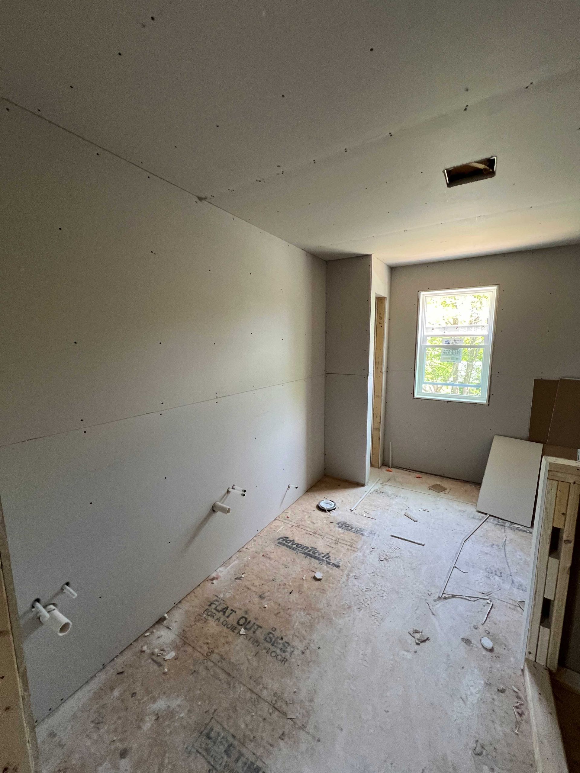 Interior room under construction; drywall walls, unfinished ceiling, window, debris on floor.