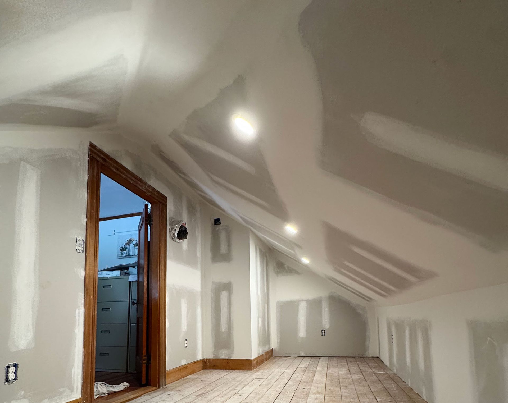 Interior of a room under construction, with drywall on walls and ceiling. Wooden trim around door and floorboards.