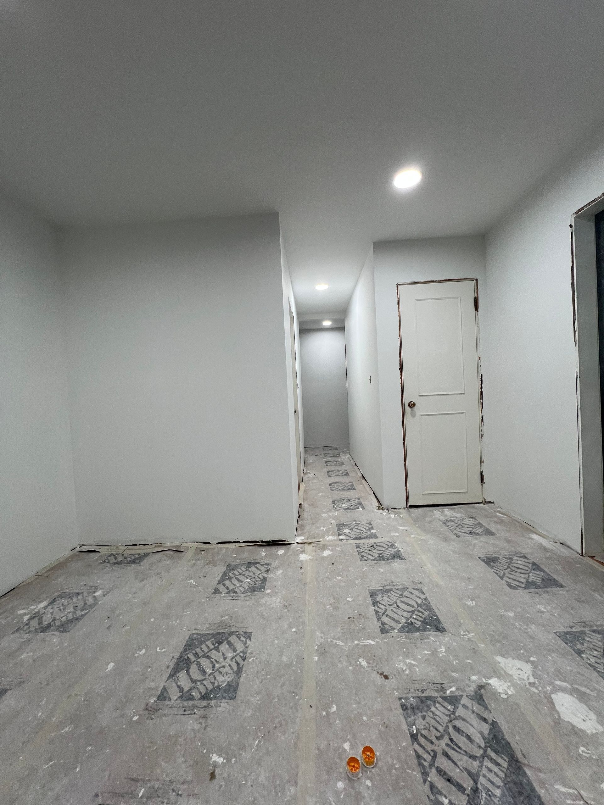 Hallway under construction, freshly painted white walls, bare floor with boot prints, closed white door, overhead lighting.