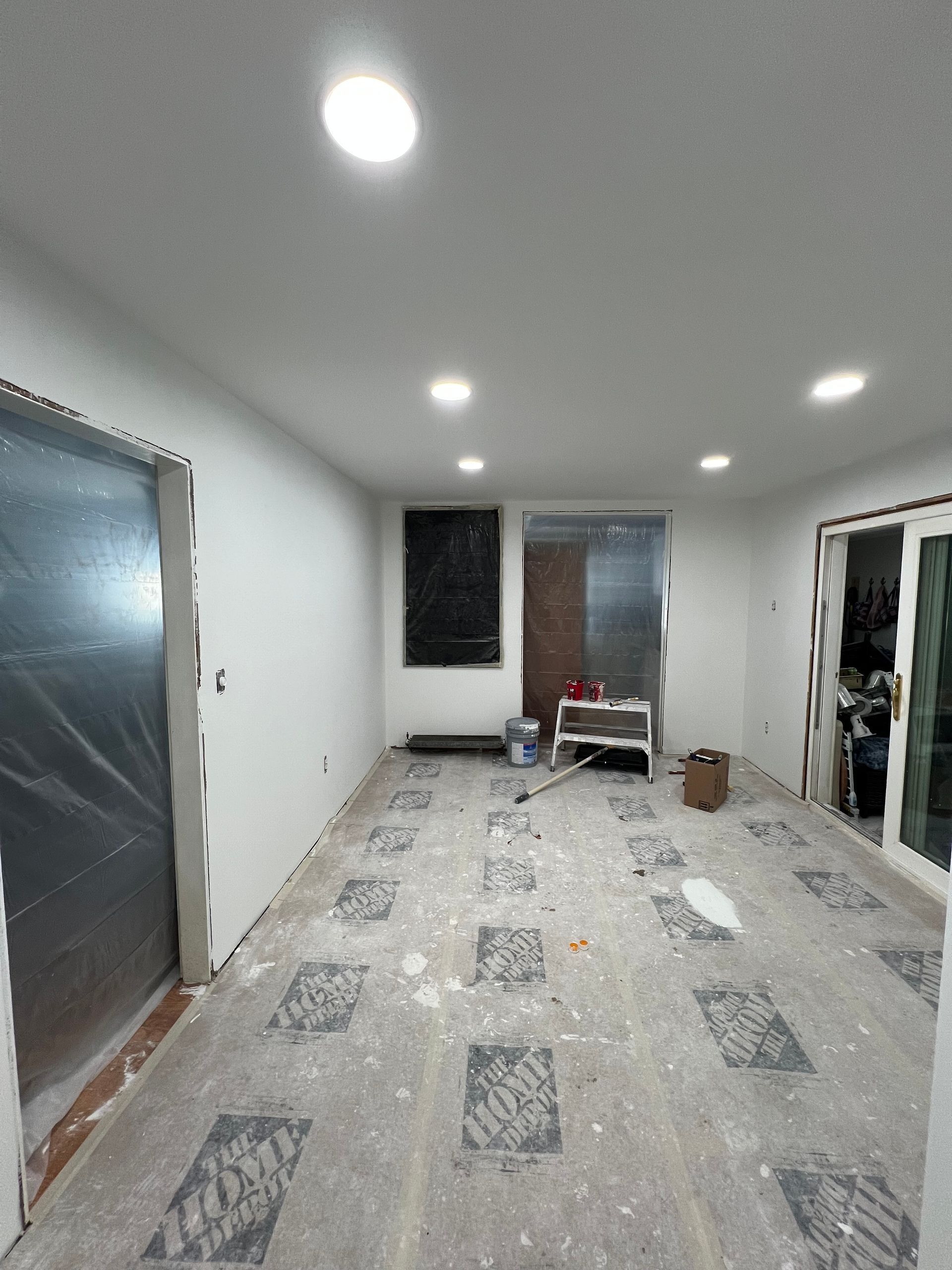Interior view of a room under renovation with white walls, ceiling lights, and protected door frames. Floor covered with protective sheeting.