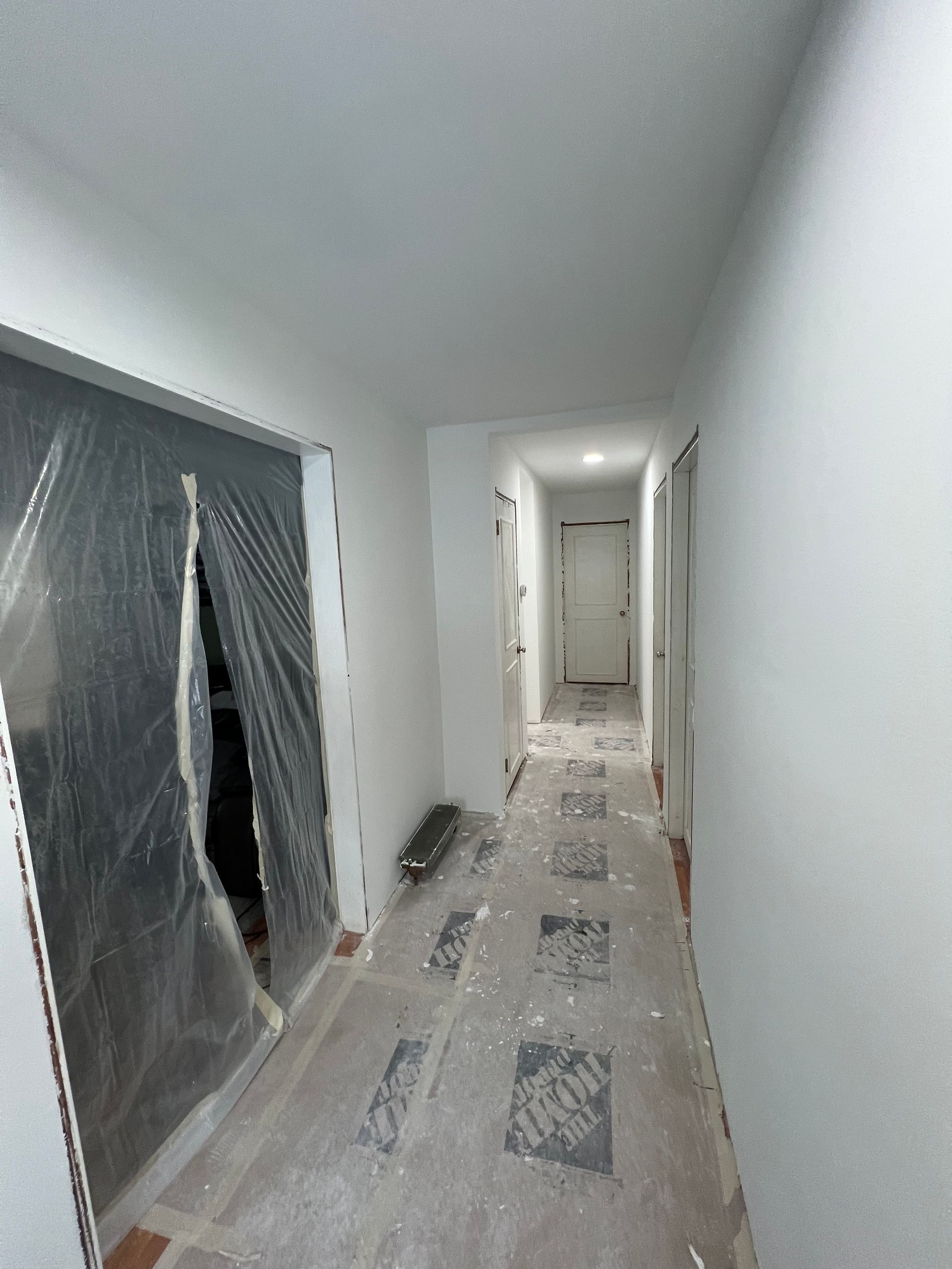 Narrow hallway, freshly painted white. Protective plastic covers a doorway. Flooring is covered with paper, debris.
