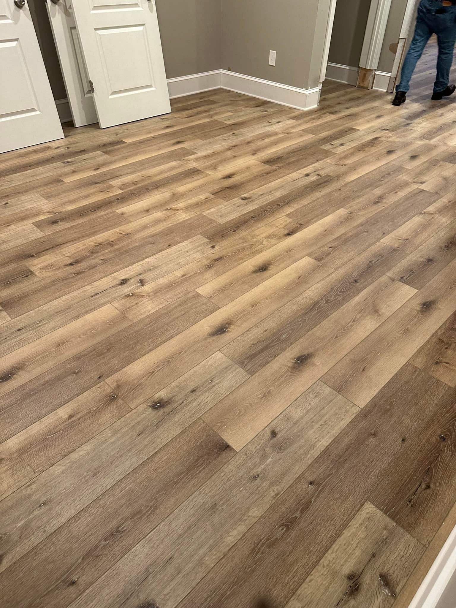 Wood-look flooring in a room with white doors and baseboards. A person's legs are visible in the background.