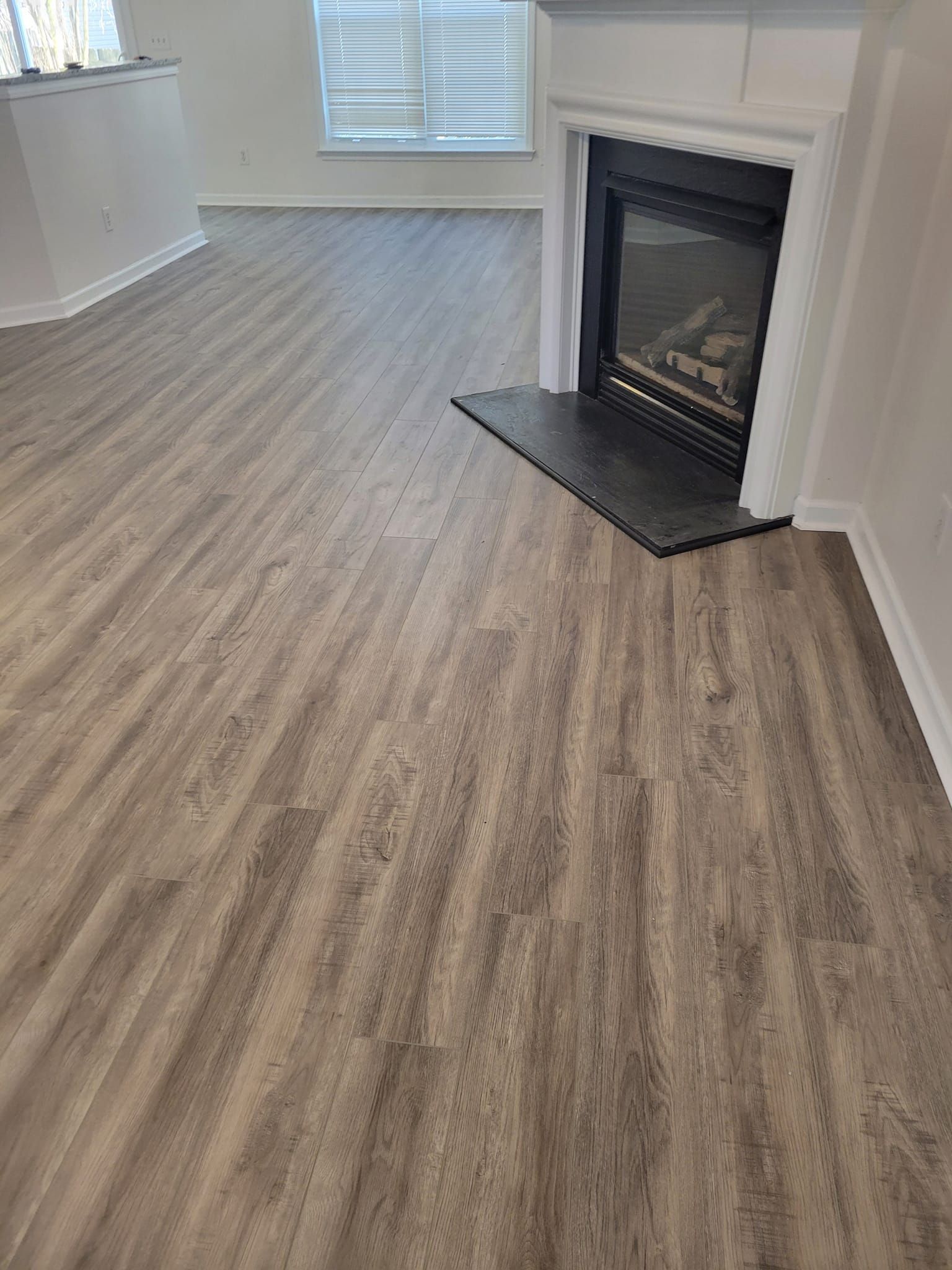 Gray wood-look flooring extends from the foreground to a fireplace with a black hearth. White trim and blinds are visible.