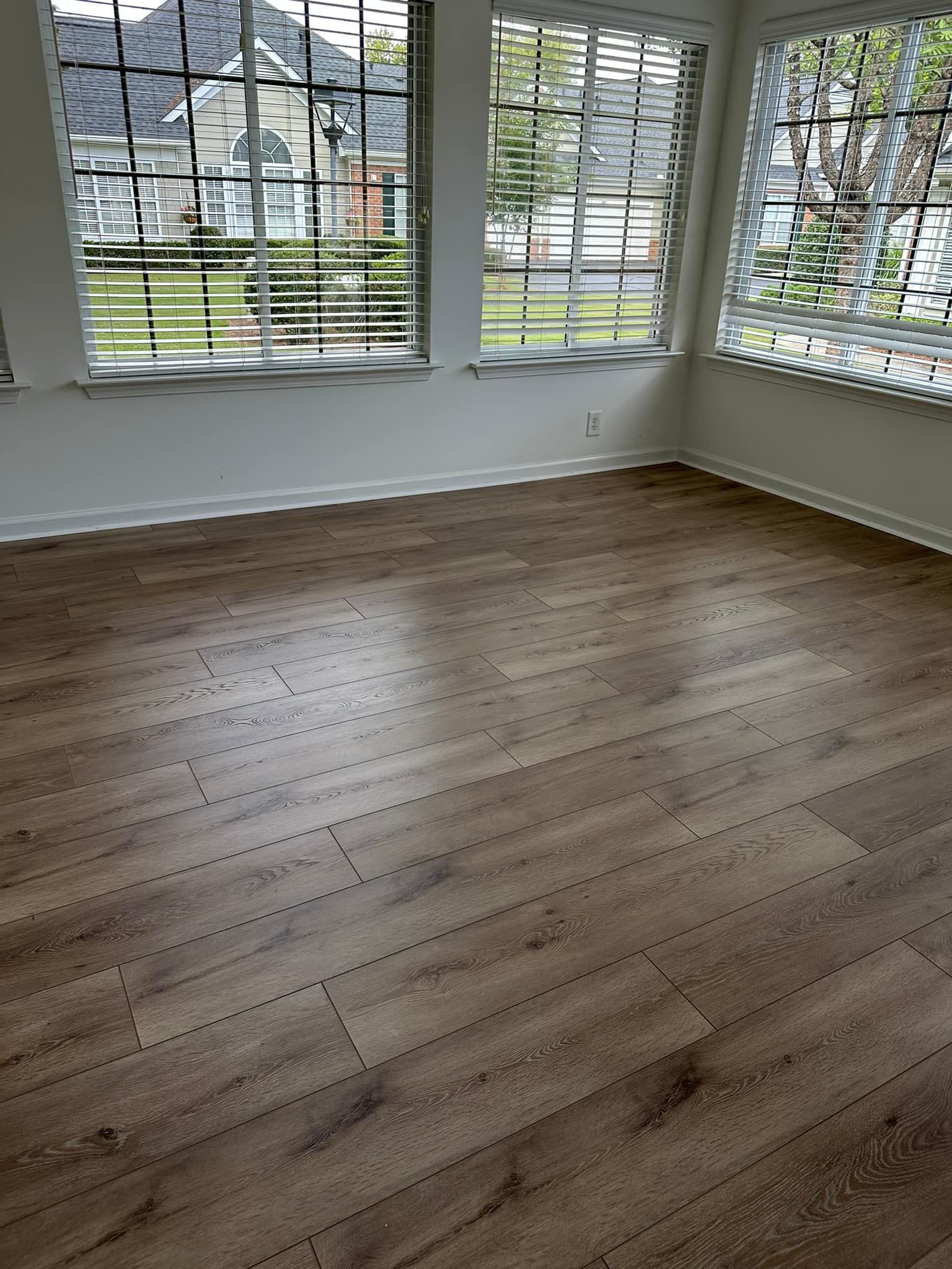 Empty room with wood-look floor, three windows with blinds, white walls, and view of houses.