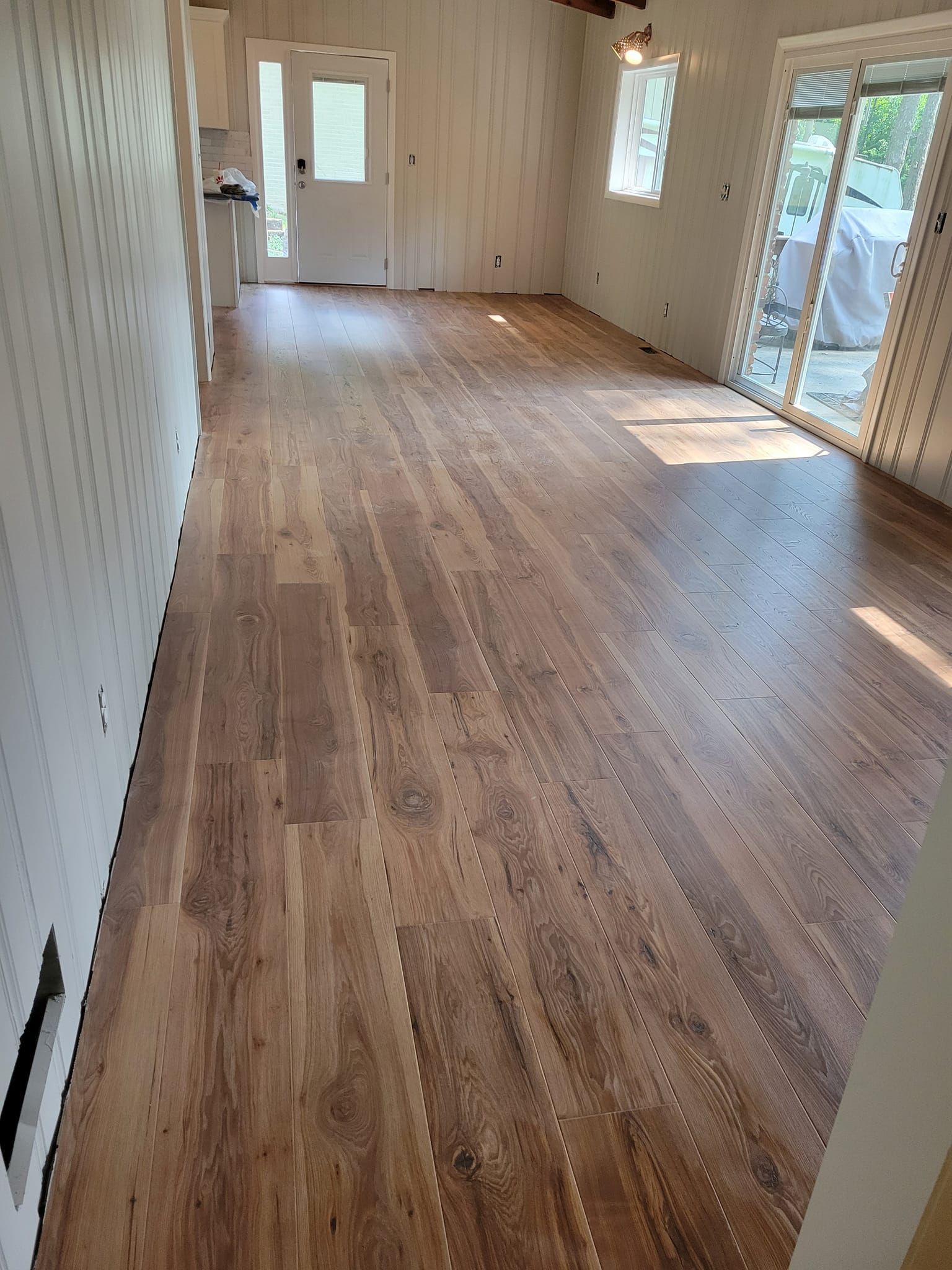 Interior view of a room with wood flooring, white walls, a sliding glass door, and a door leading to a kitchen.