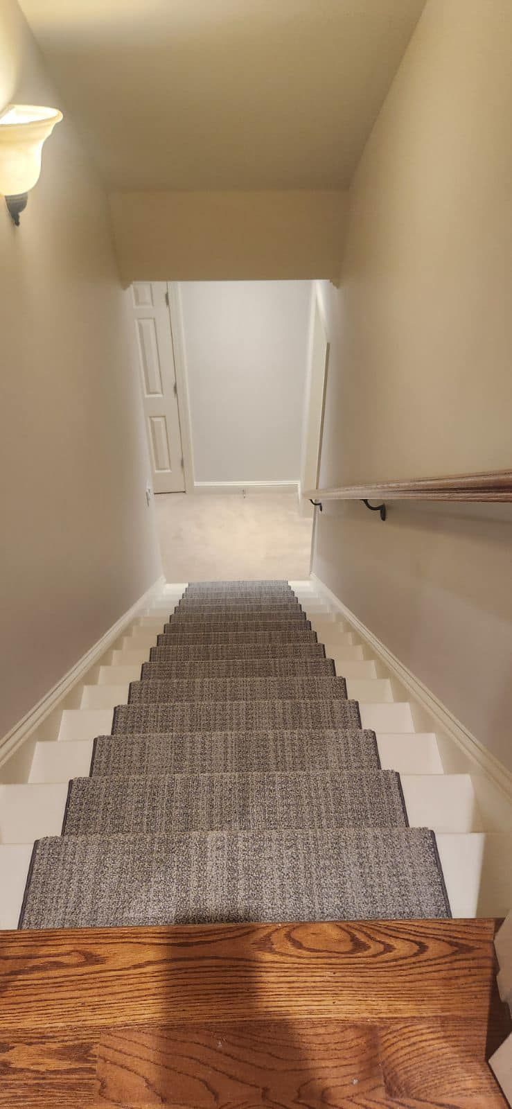 Staircase descending into a hallway with carpeted steps. Beige walls, wooden handrail, and a door at the bottom.