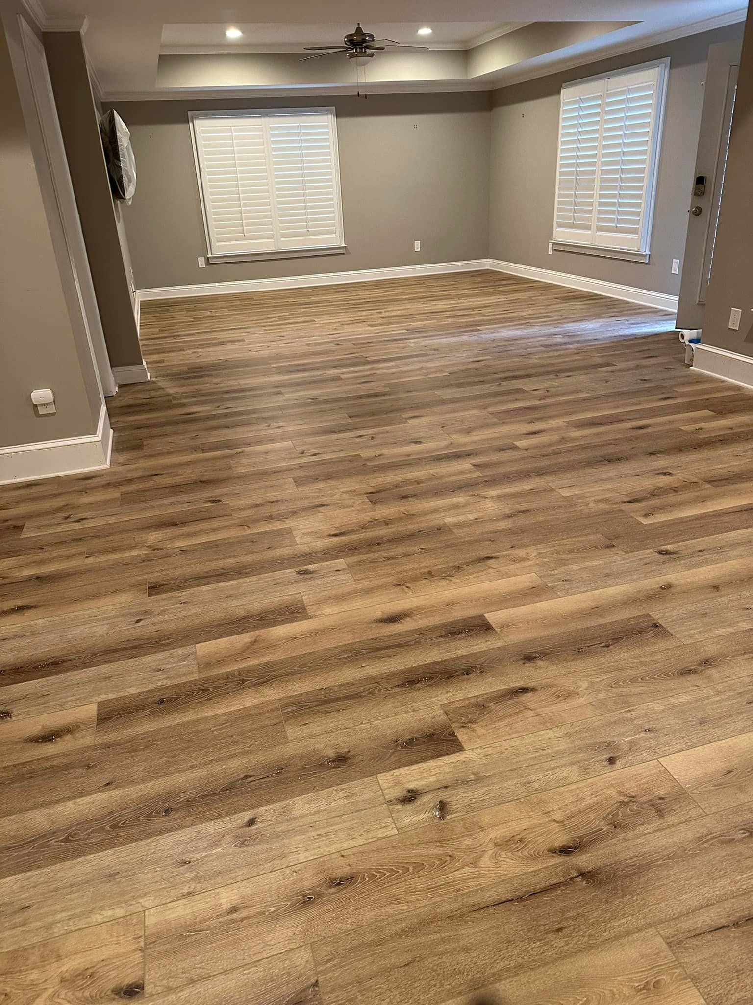 Empty room with light brown wood-look flooring, beige walls, and white trim. Two windows with blinds are visible.