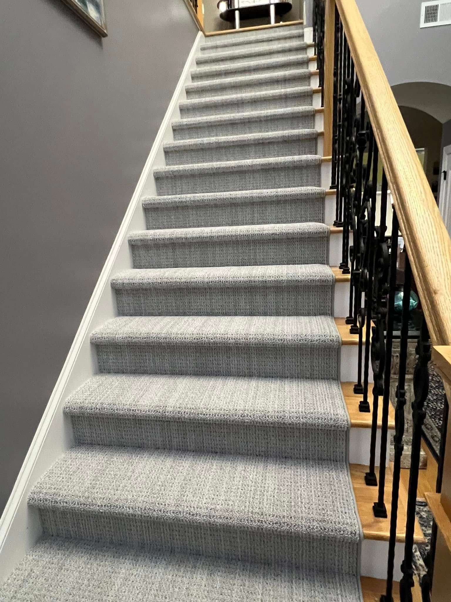 Staircase with gray carpeted steps, white risers, wooden handrail, and black wrought iron balusters.