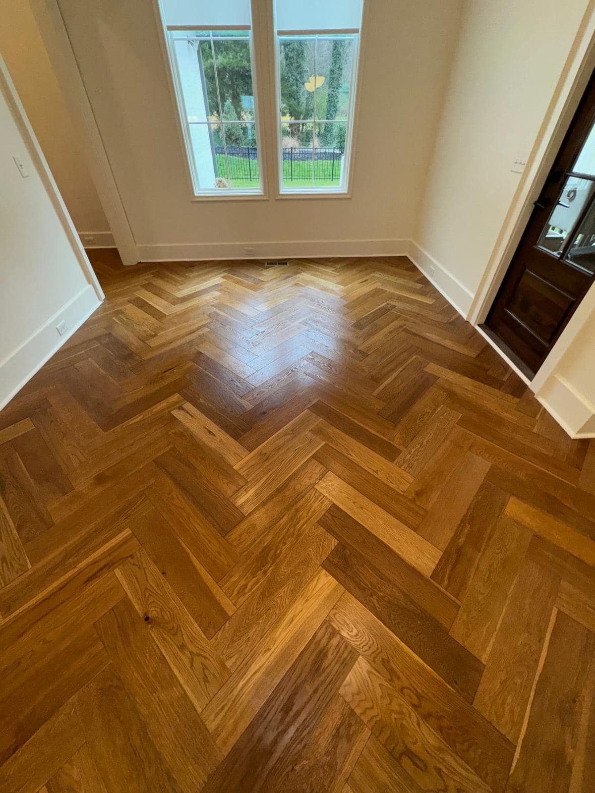 Herringbone pattern hardwood floor in a room with a window and a dark wooden door.