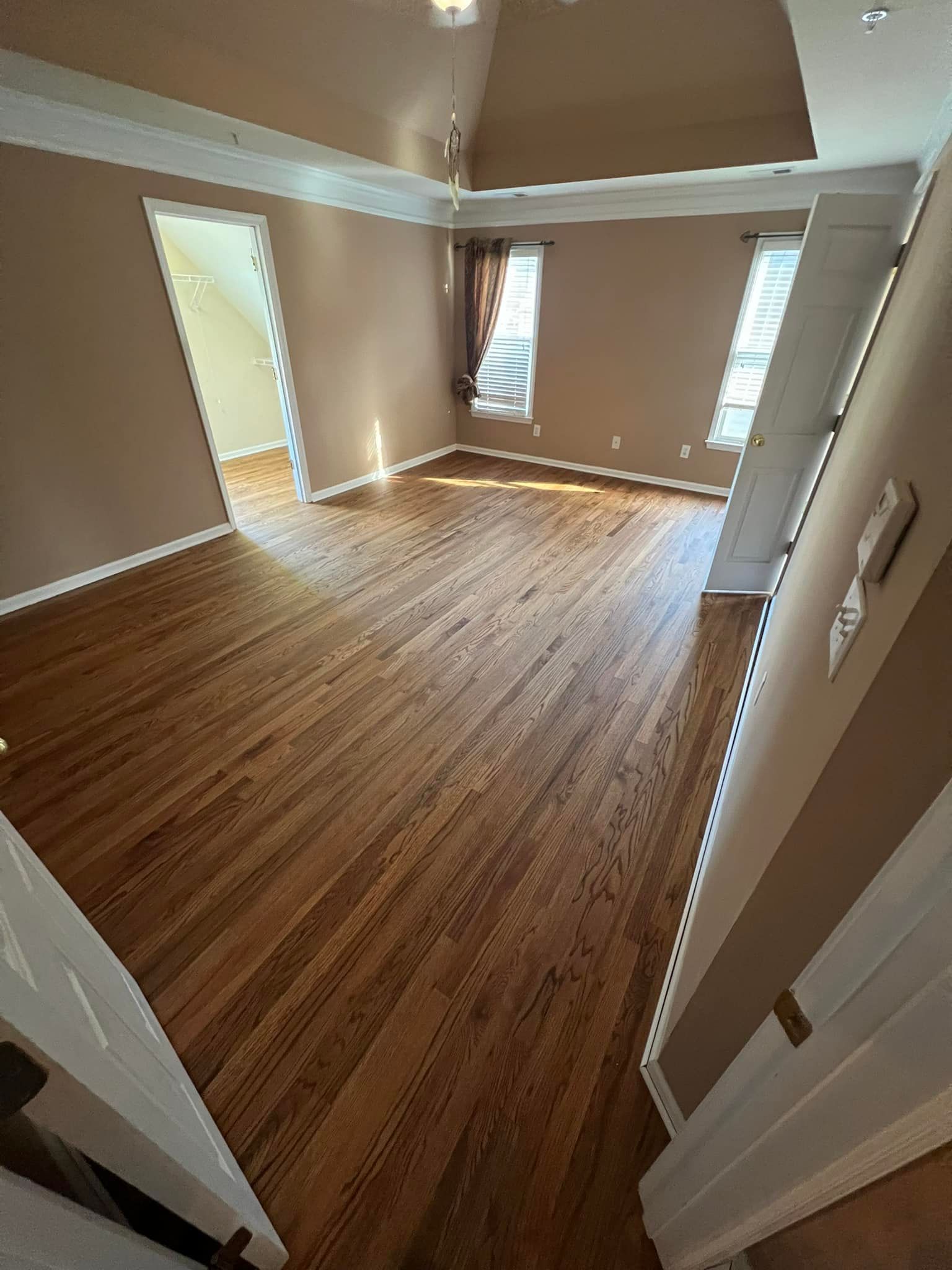 Hardwood floors in a room with tan walls and white trim. Sunlight streams in from a window.