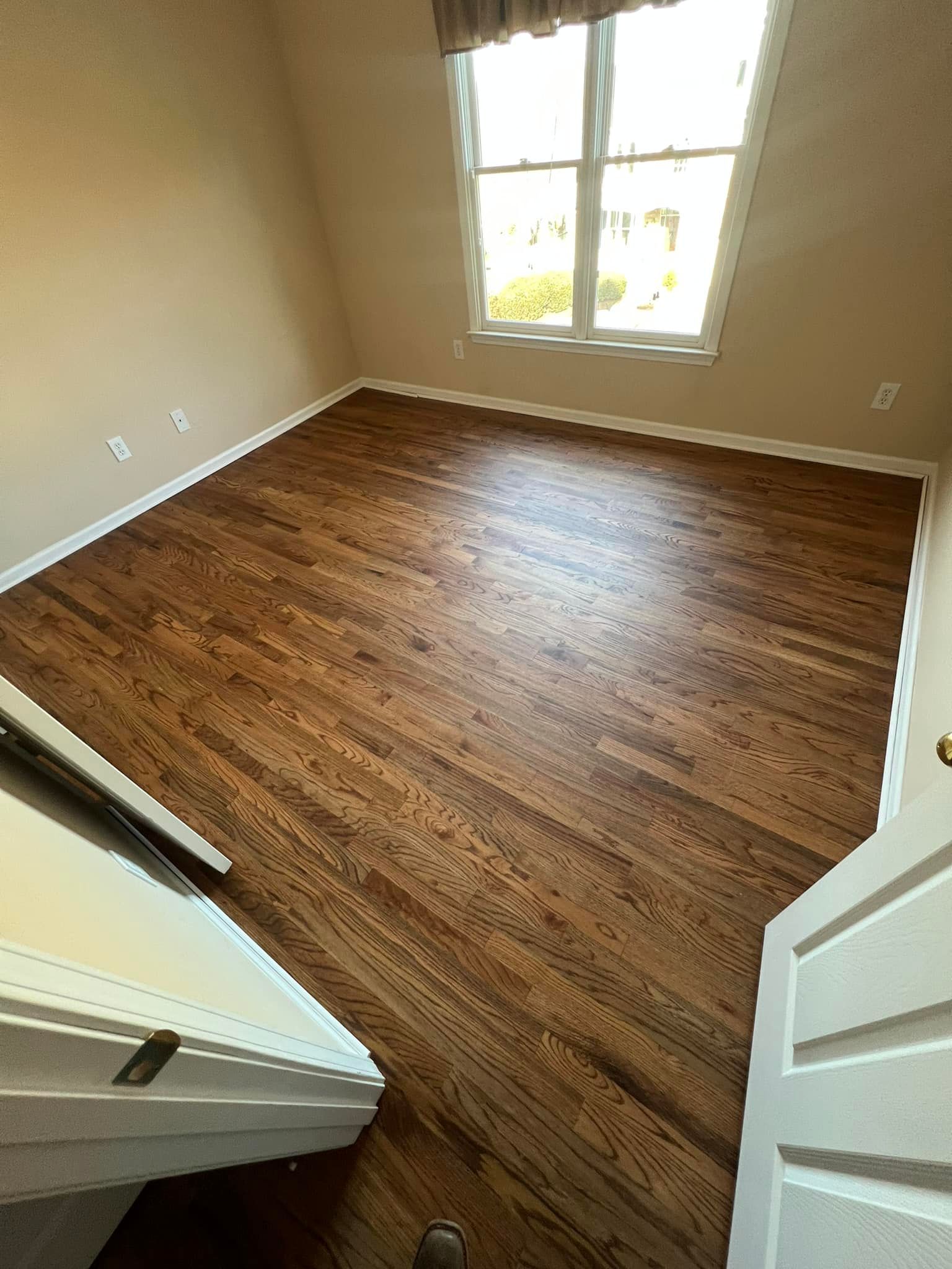 Hardwood floor in room with beige walls and a window with blinds. Open white door in foreground.