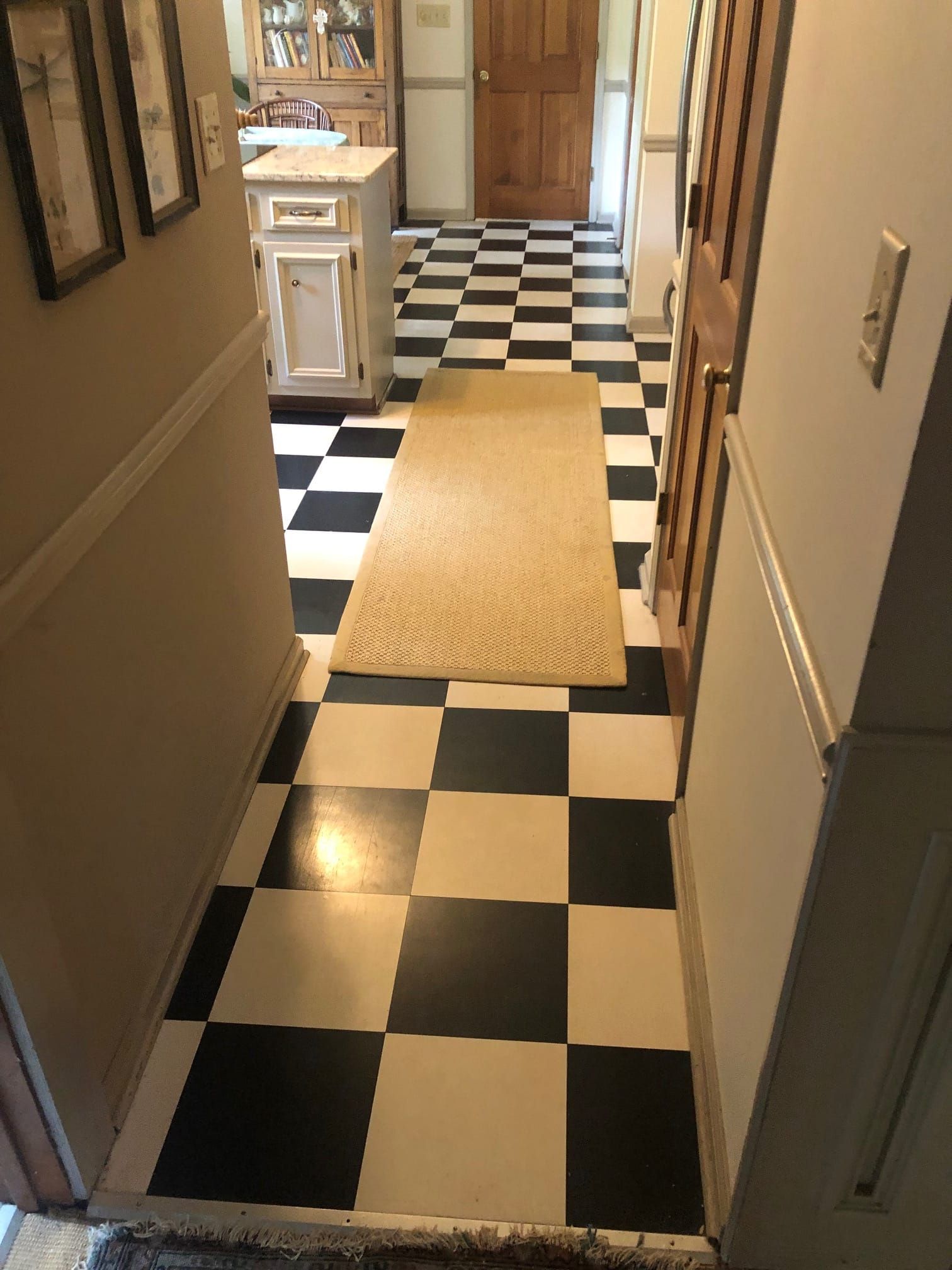 Hallway with black and white checkered floor, beige rug, and beige walls, leading to a wooden door.