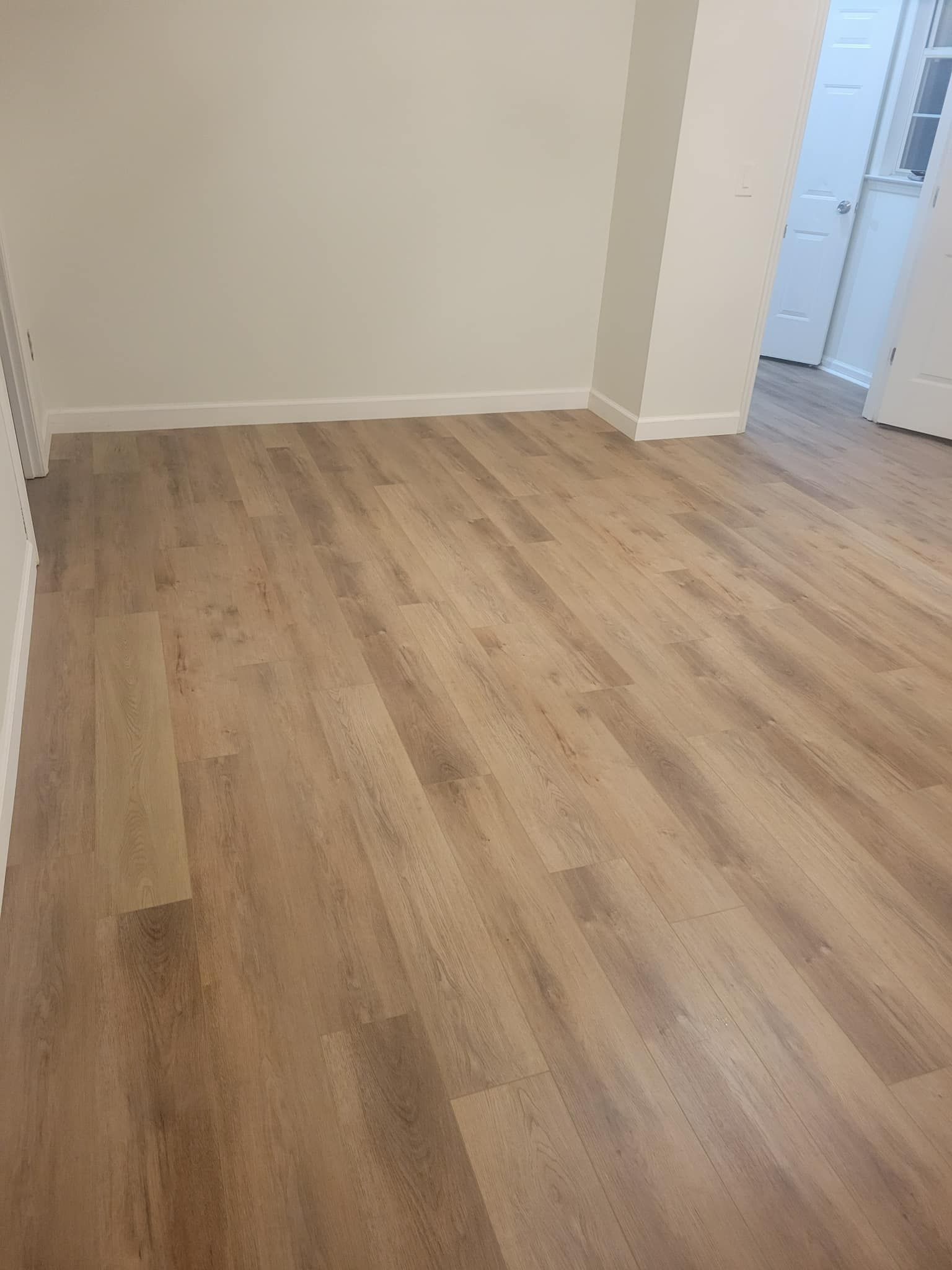 Empty room with light wood-look flooring and white walls, door in the distance.