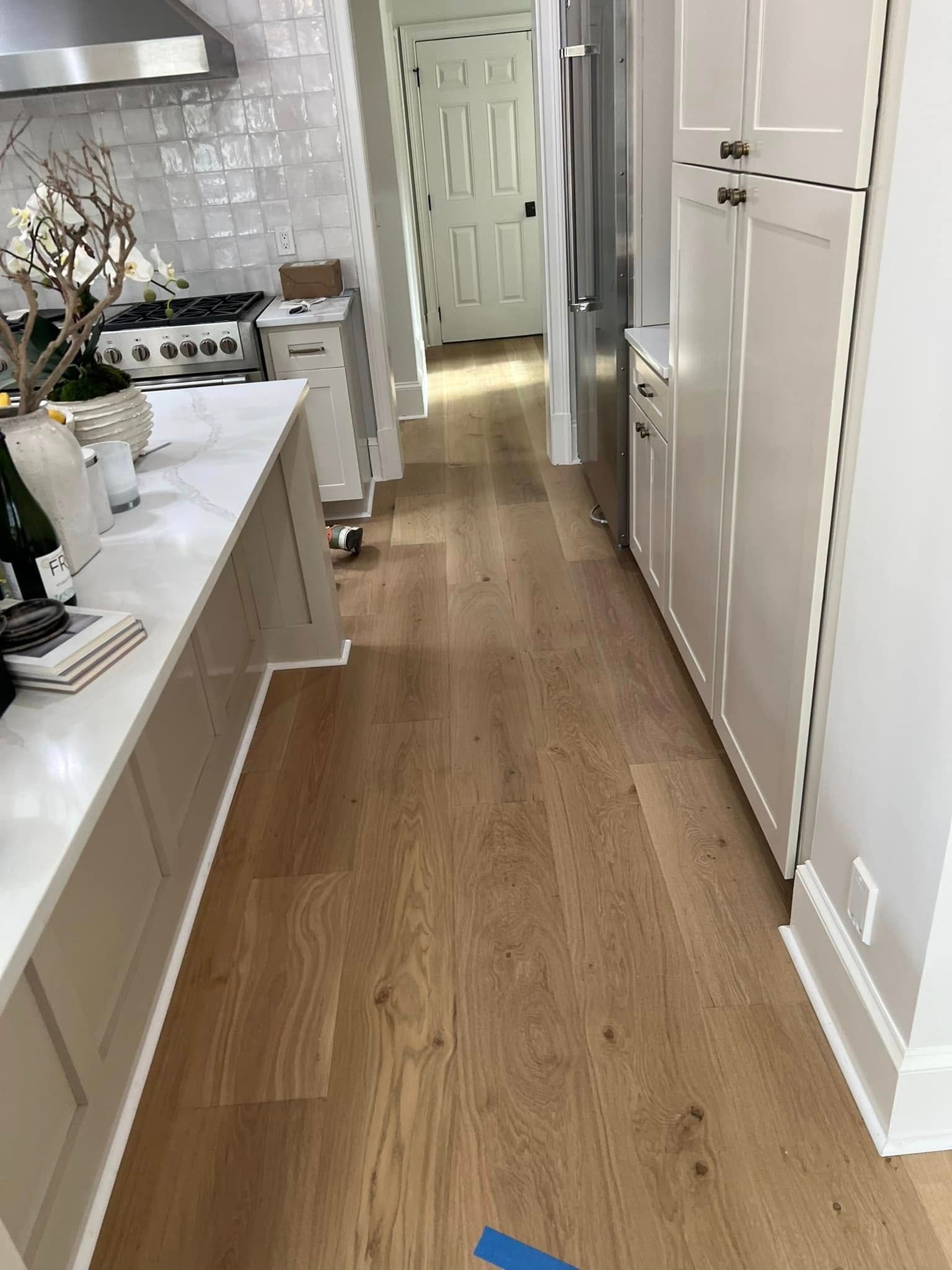 Wooden floor in a kitchen with white cabinets, a long countertop, and a stainless steel fridge.