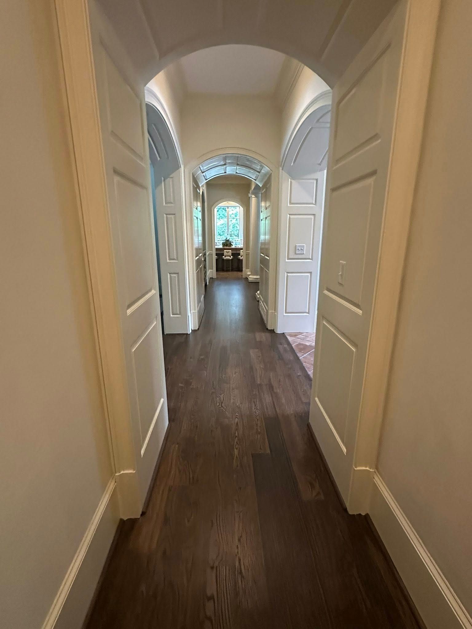 Narrow hallway with dark wood floor and arched doorways leading to a room with a window.