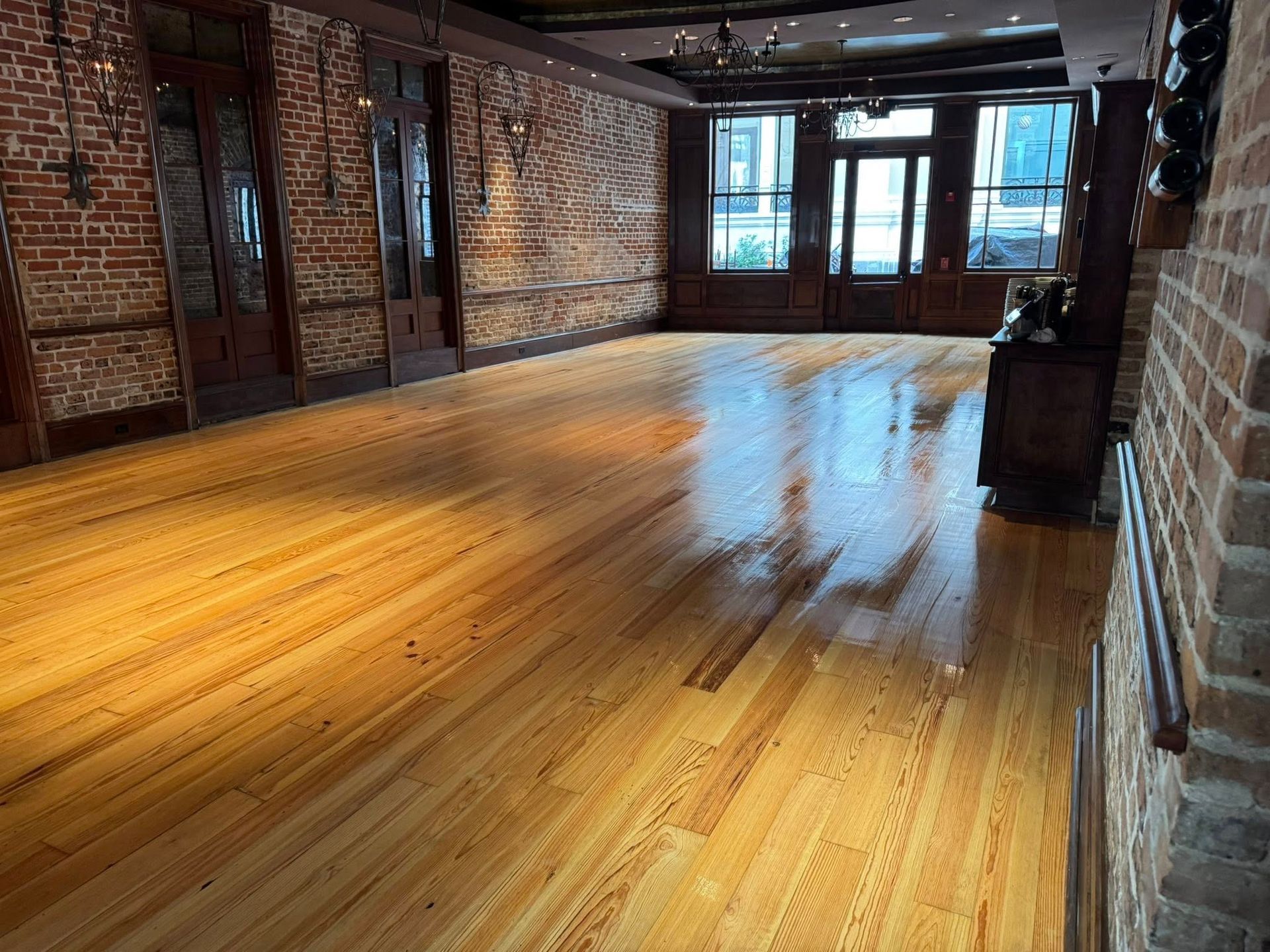 Empty room with wooden floor and brick walls, large windows in the background.