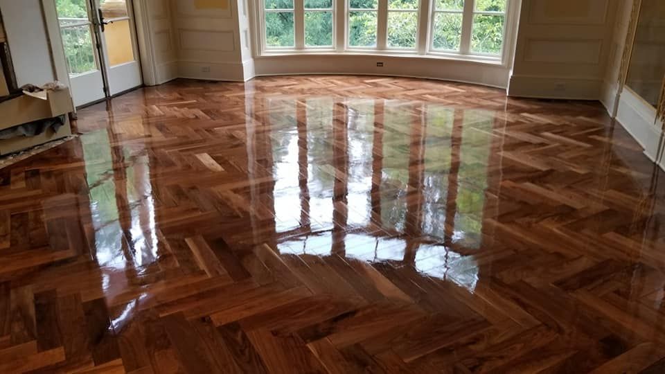 Shiny, newly-polished parquet flooring in a room. Reflections of a window and trees are visible on the floor.