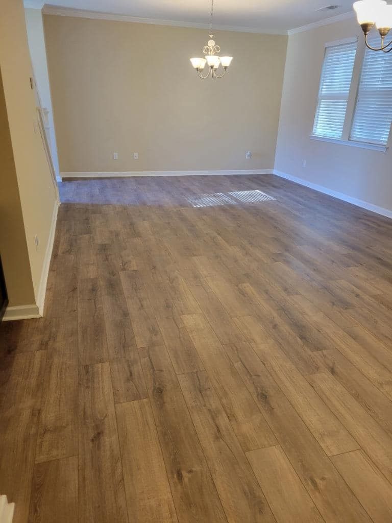 Empty living room with wood-look flooring, beige walls, chandelier, and a window with blinds.