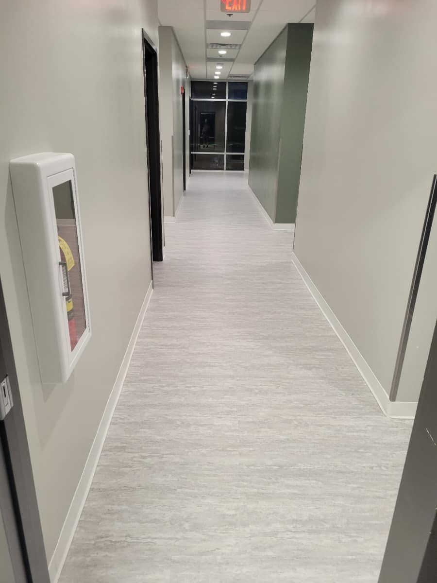 Long, narrow hallway with light gray flooring and pale walls. A fire extinguisher box is on one wall.