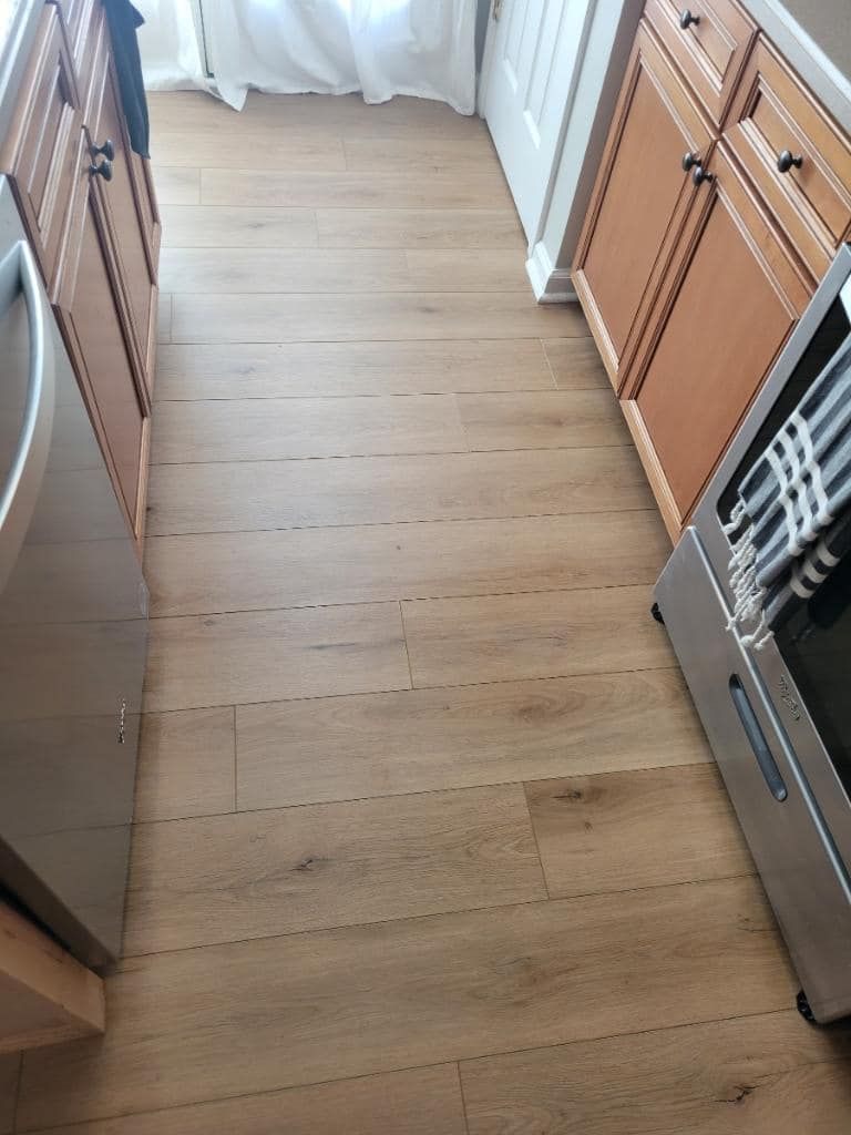 Wooden-floored kitchen, with cabinets, stainless steel appliances, and a window with white curtains.