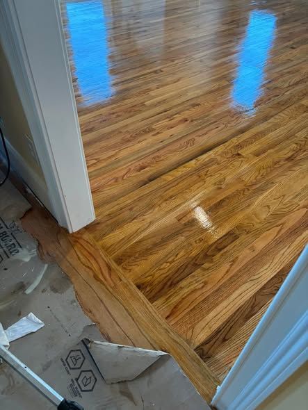 Refinished hardwood floors, transitioning between rooms, reflecting sunlight. Edge detail and unfinished area visible.