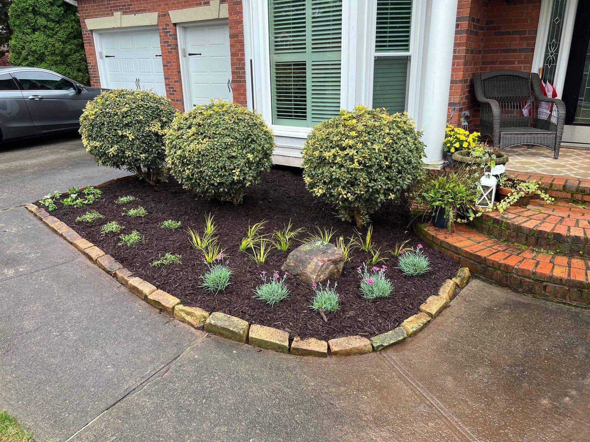 A garden in front of a brick house with a car parked in the driveway.