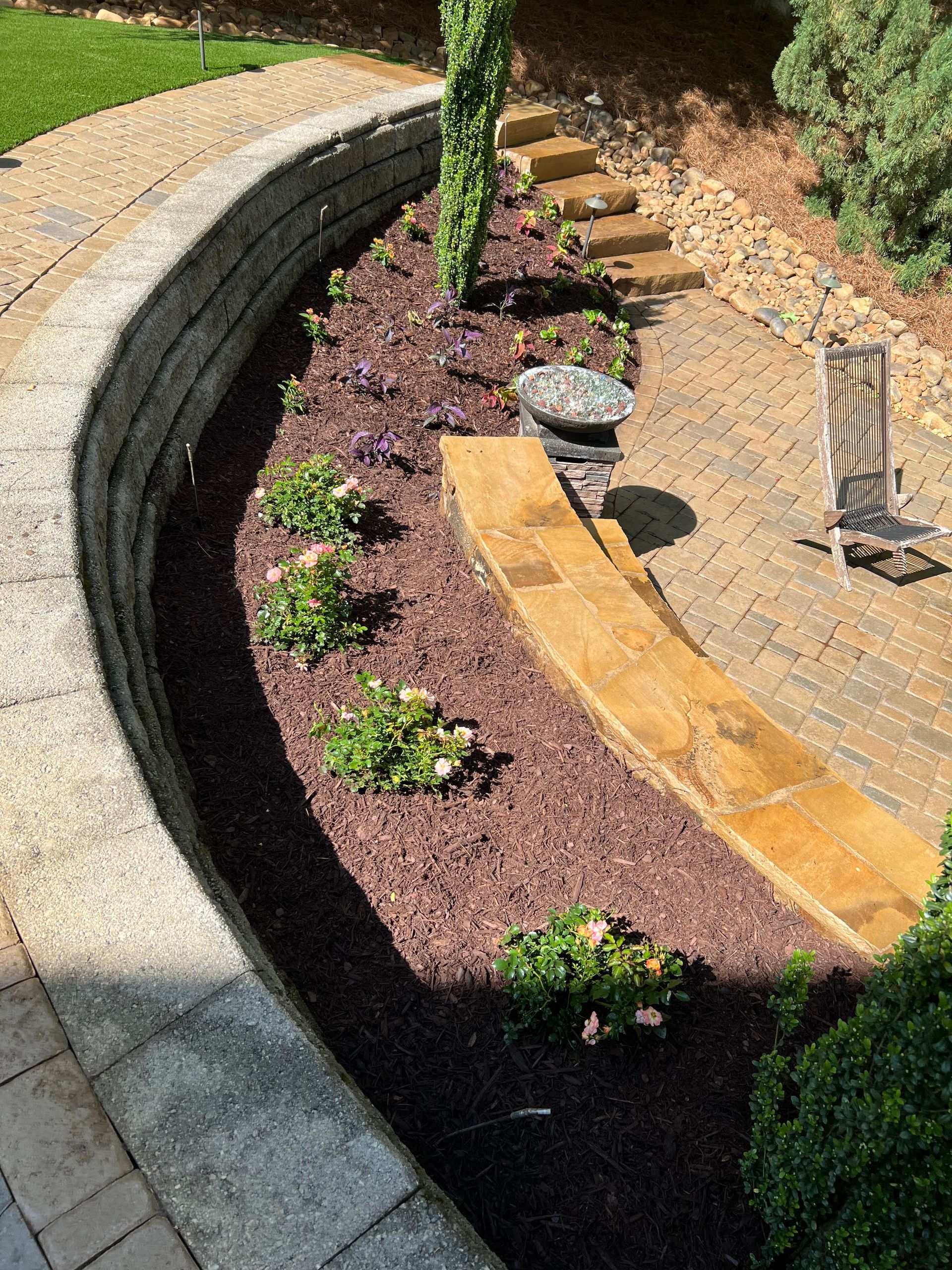 A brick walkway leading to a patio with a stone wall and a chair.
