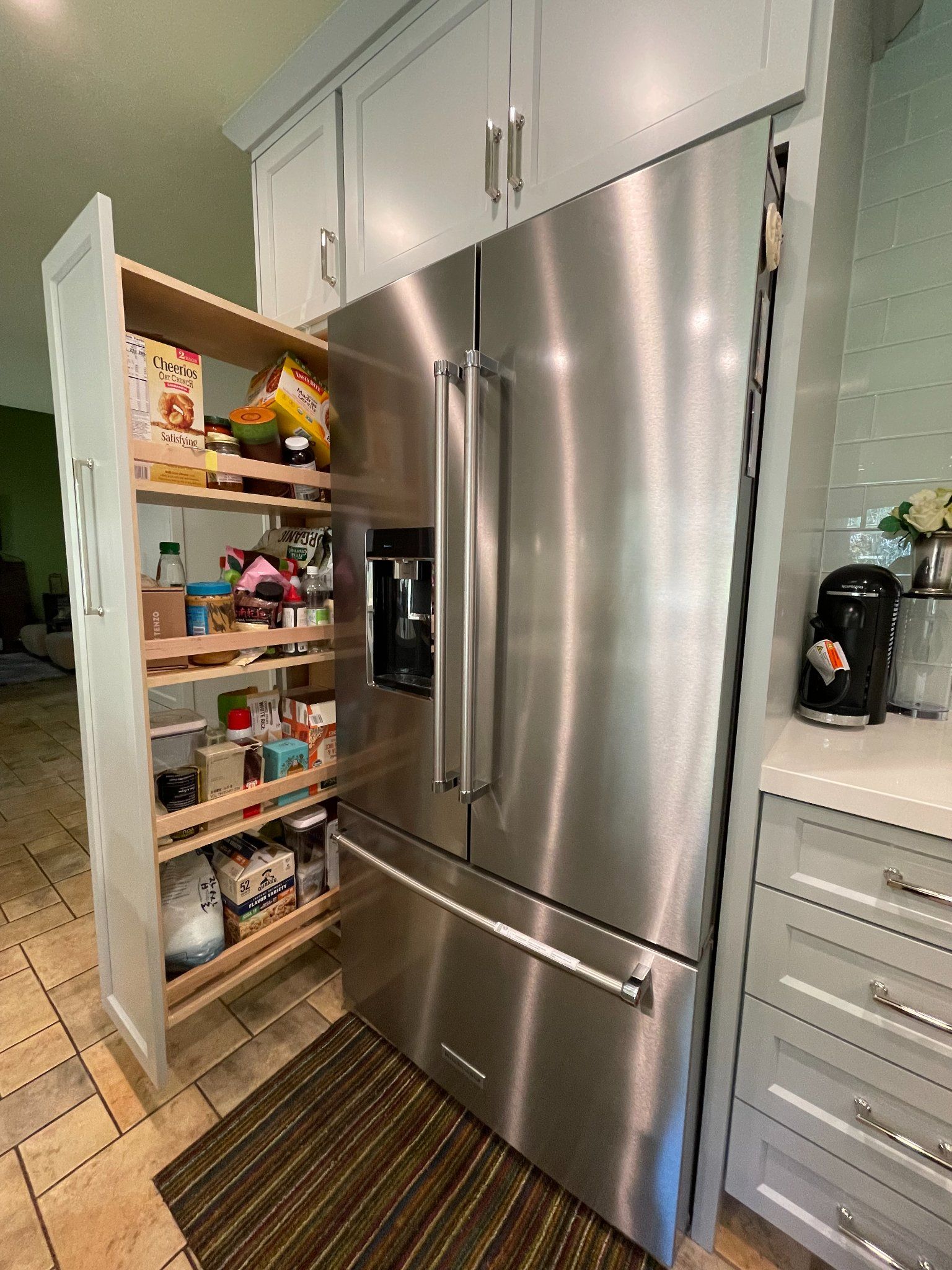Pull-out pantry next to a stainless steel refrigerator in a kitchen with light-colored cabinets and tile flooring.