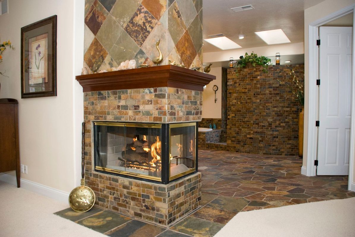 Corner fireplace with glass sides, tiled surround, and wooden mantel in a room with stone flooring.