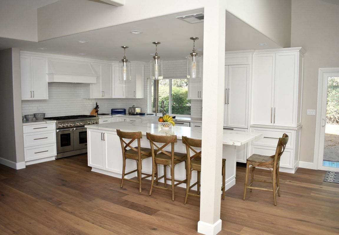White kitchen with island, stainless steel appliances, and wooden flooring. Four brown stools at island.