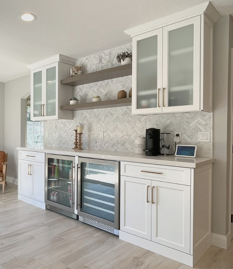 White built-in bar with cabinets, beverage coolers, open shelves, and a coffee machine against a tiled wall.