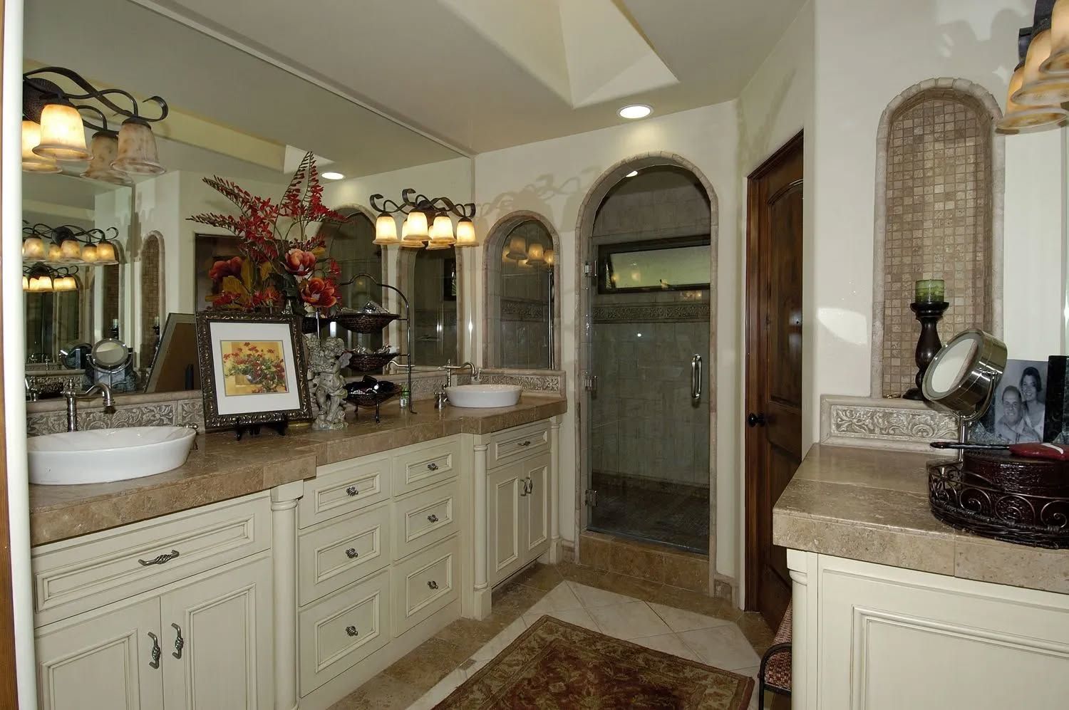 Bathroom with beige cabinets, light-colored countertops, large mirrors, and an arched entry to a shower.