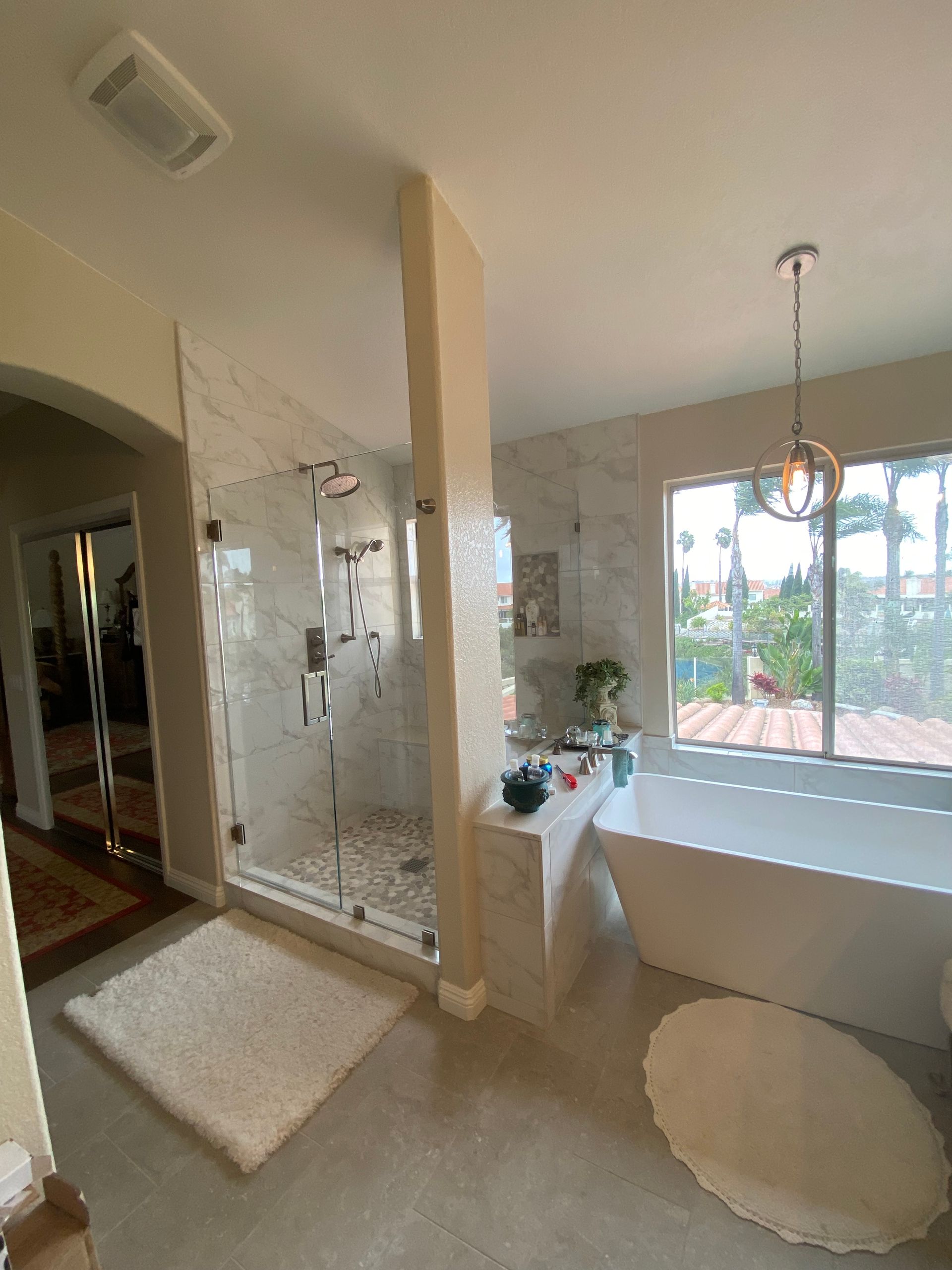 Modern bathroom with glass shower, bathtub, large window, and tan walls.
