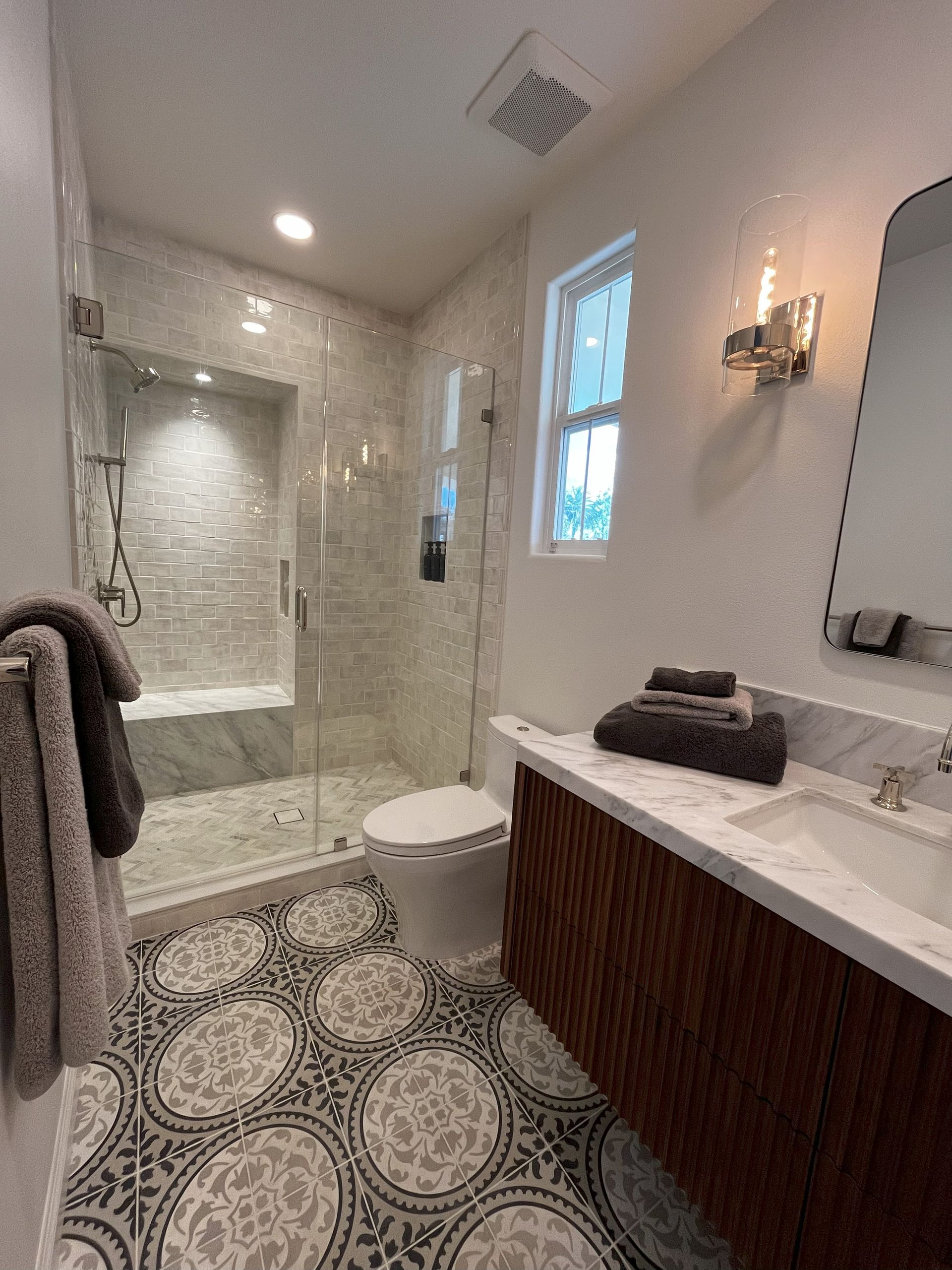 Bathroom with patterned tile floor, glass shower, wooden vanity, and marble countertop.