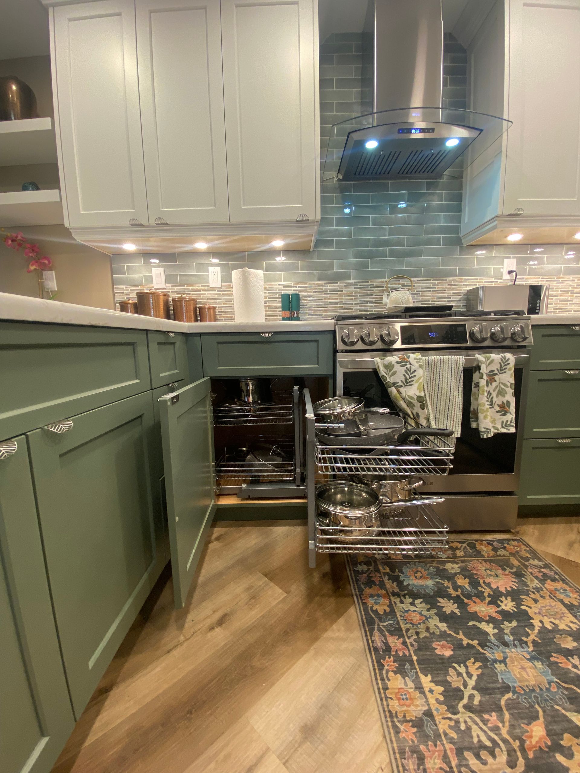 Green kitchen with pull-out cabinet. Stainless steel range and hood, backsplash, wood floor, and rug.