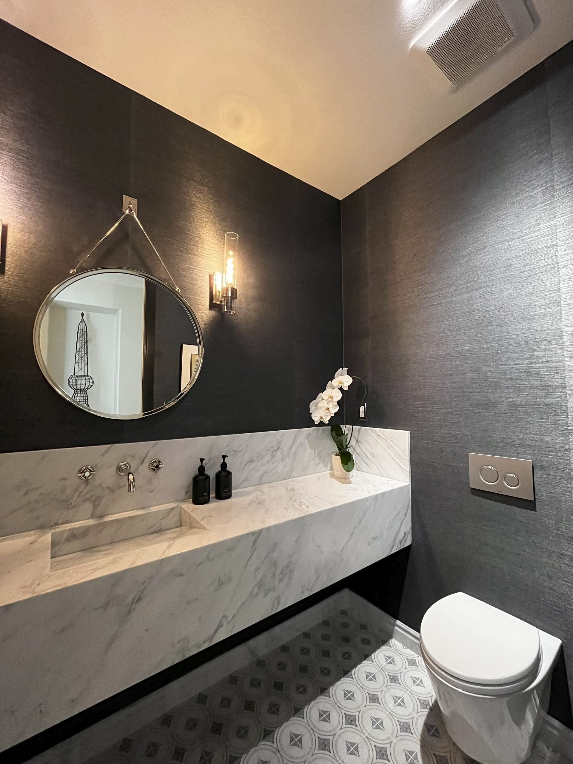 Modern bathroom with dark textured walls, marble countertop, round mirror, and patterned floor.