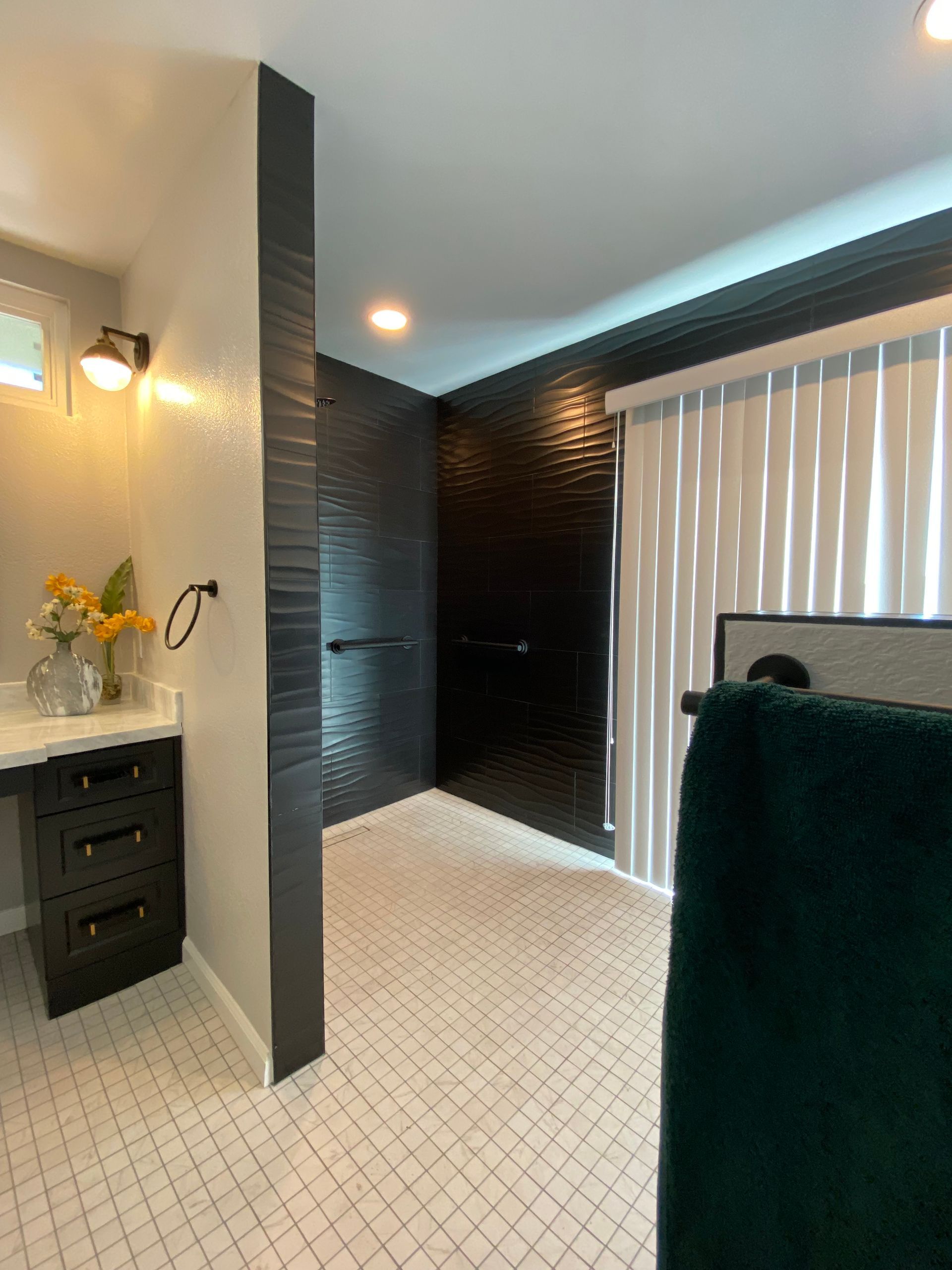 Modern bathroom with black tiled shower, white tile floor, dark vanity, and sheer window coverings.