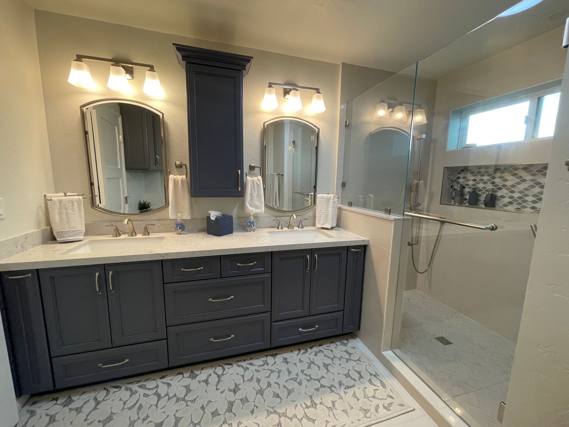Bathroom with blue cabinets, marble countertop, glass shower, and two mirrors.