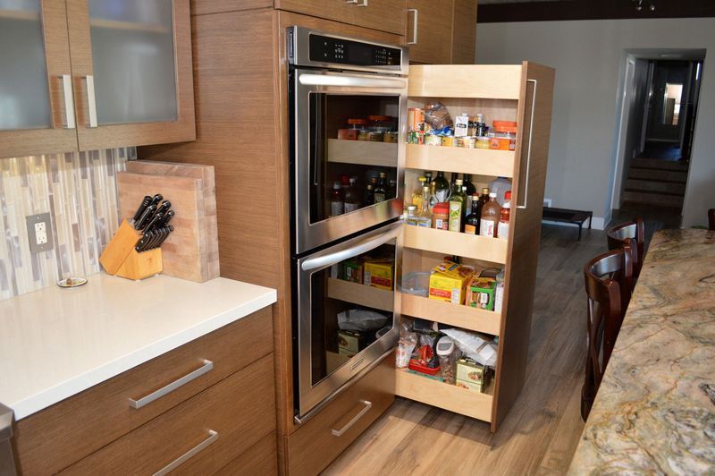 Modern kitchen with pull-out pantry next to a double oven. Wooden cabinets, white countertop, tile backsplash.