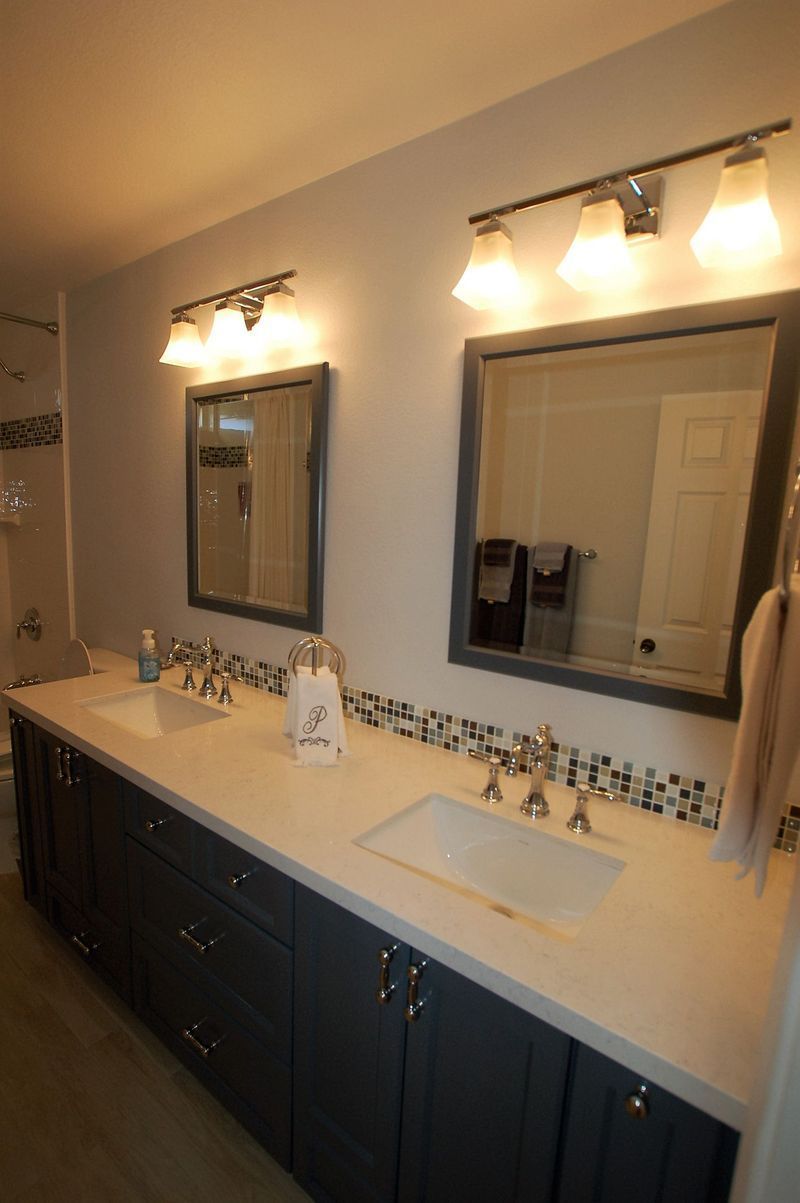 Bathroom vanity with two sinks, mirrors, and lights; dark blue cabinets, light countertop, mosaic tile accent.