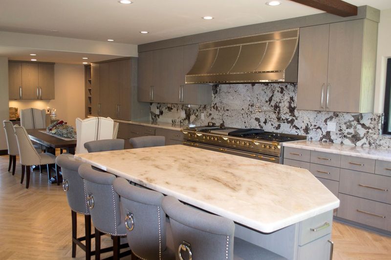 Modern kitchen with gray cabinets, island seating, stainless steel range and hood, and marble backsplash.